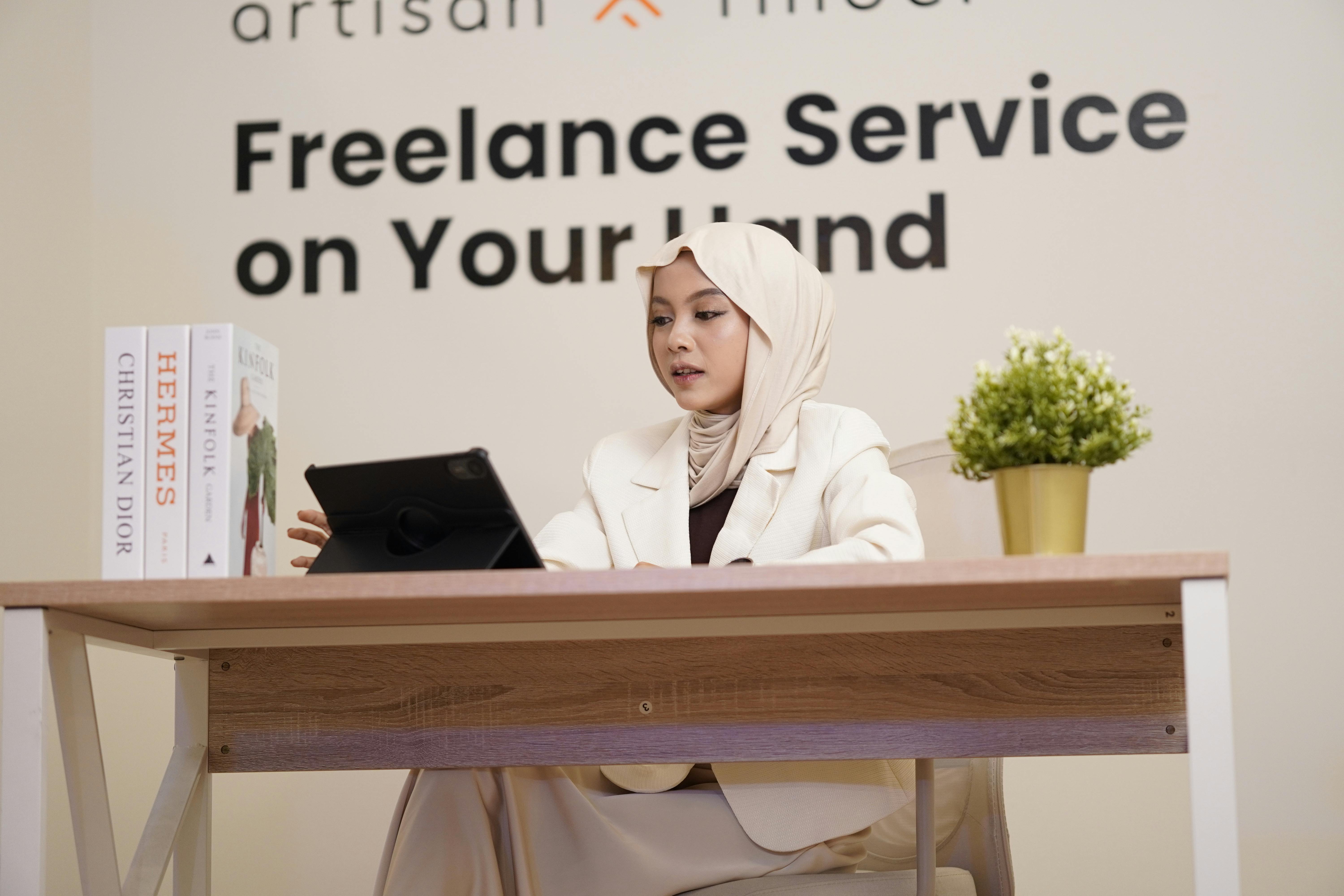 Professional woman in hijab working on a tablet at a modern office desk with books and decor.