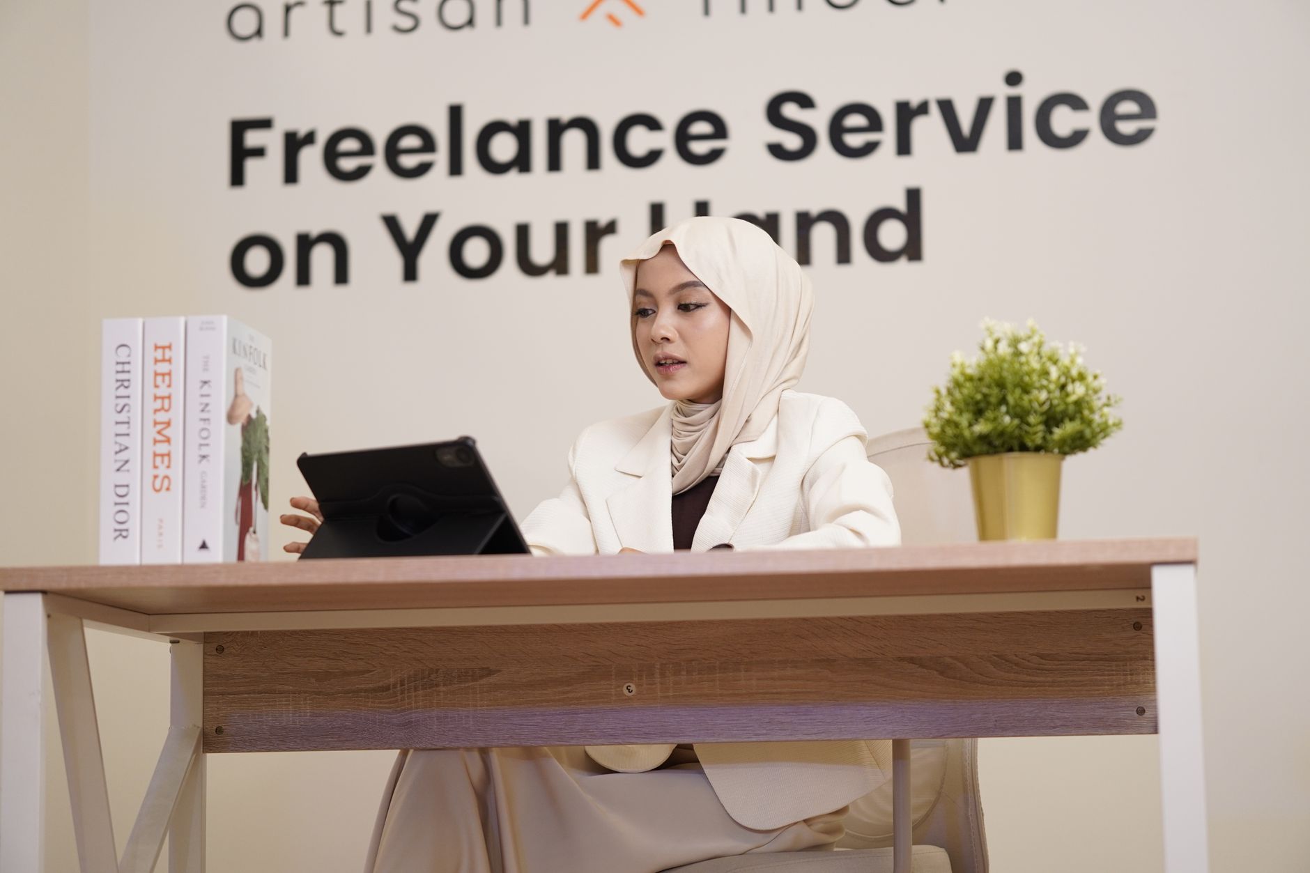 Professional woman in hijab working on a tablet at a modern office desk with books and decor.