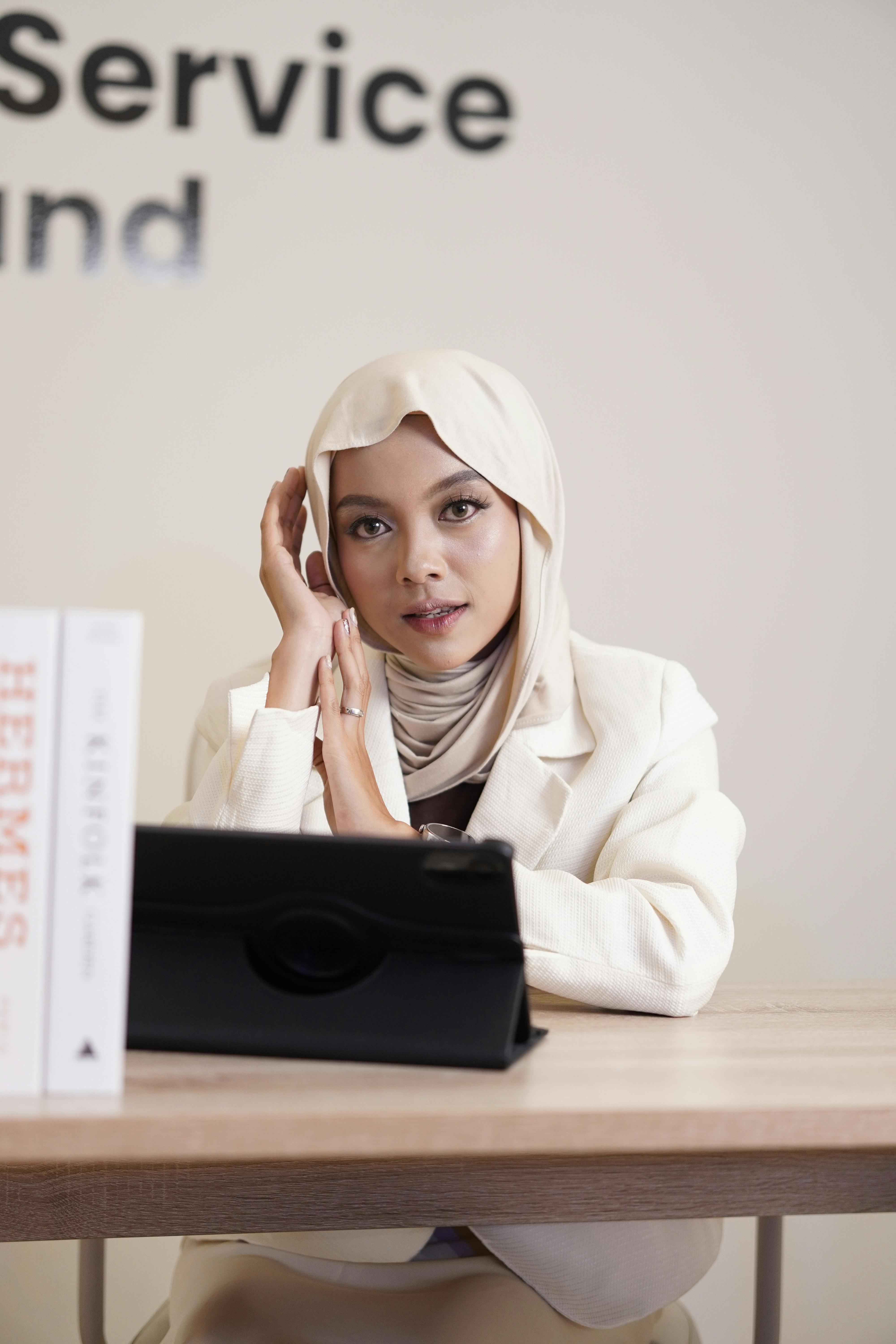 Confident businesswoman in hijab working at a desk with tablet and books in modern office.