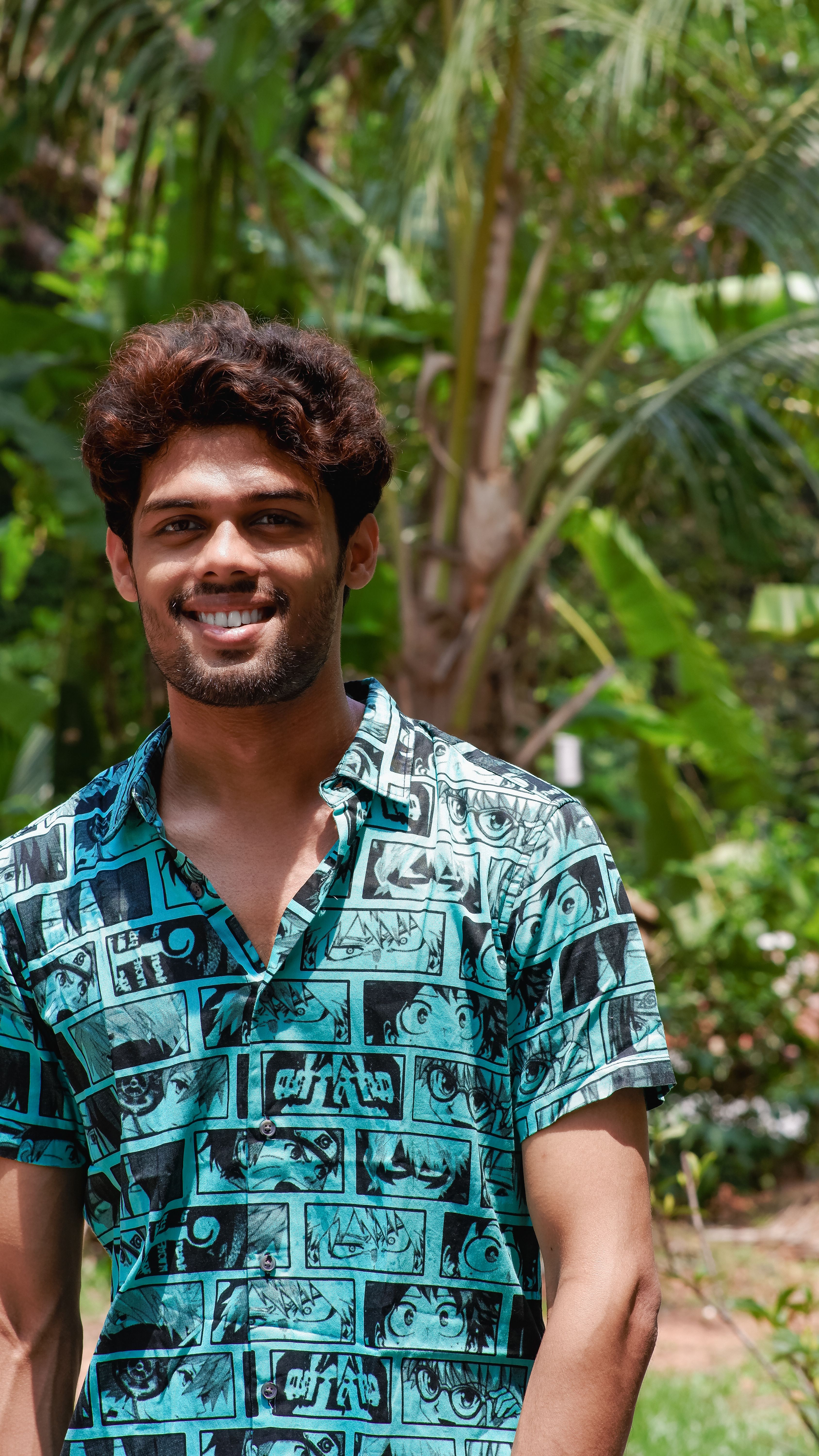 A man smiles in a lush garden, wearing a modern patterned shirt.