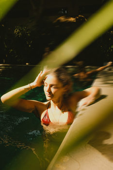 A woman in a red bikini enjoys sunlight by a pool, surrounded by greenery.