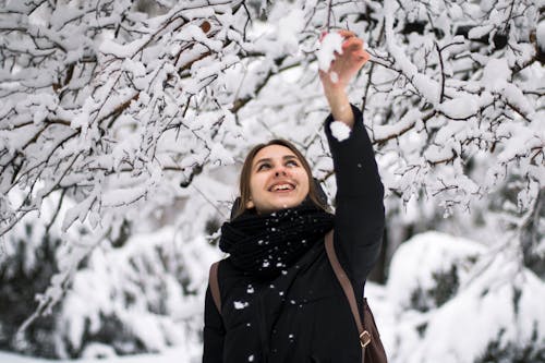 Фотография улыбающейся женщины в черном зимнем пальто, стоящей под заснеженным деревом