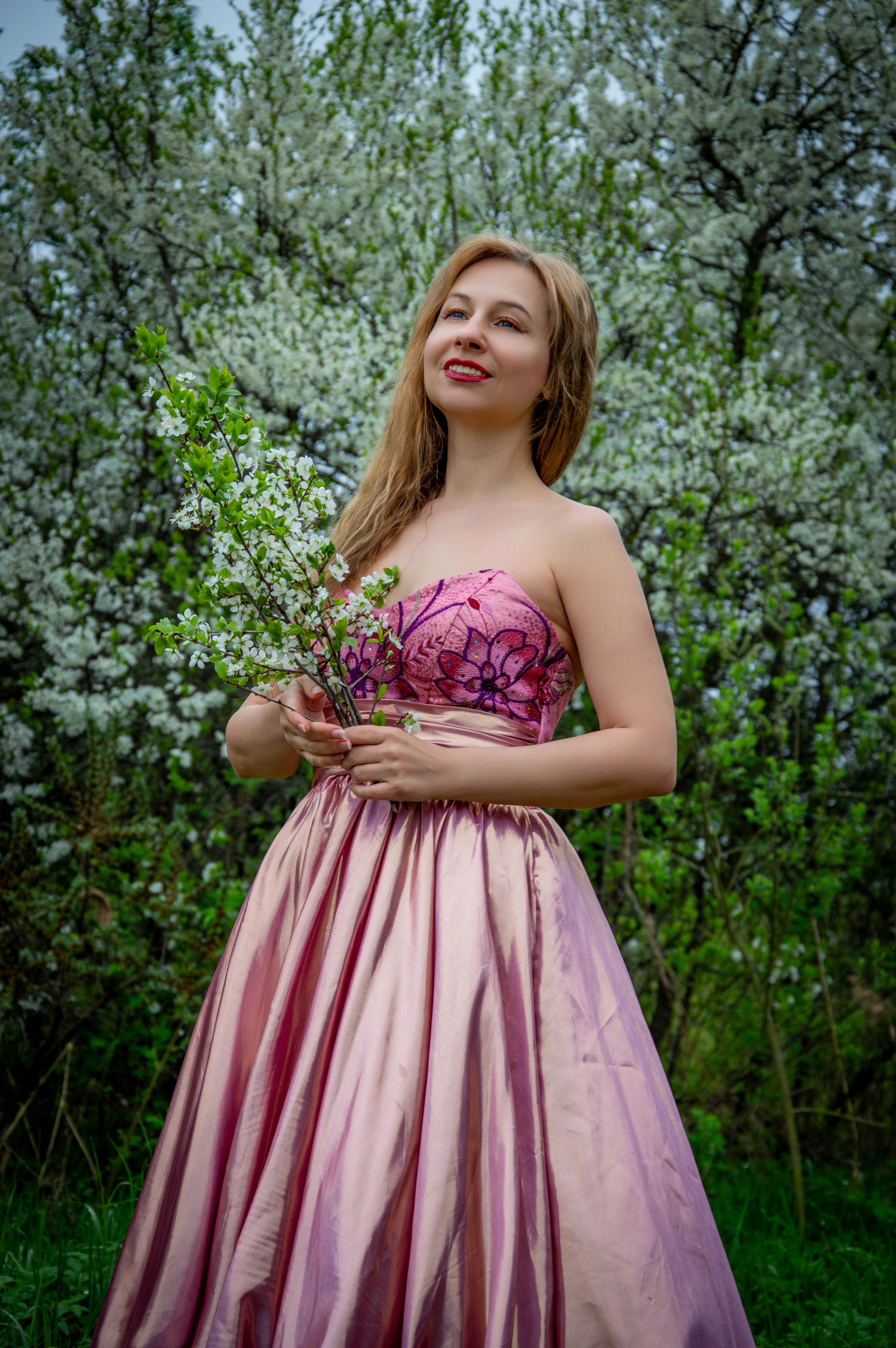 A woman in a pink gown holding white blossoms stands in a flowering garden during spring. Captured with vibrant detail.