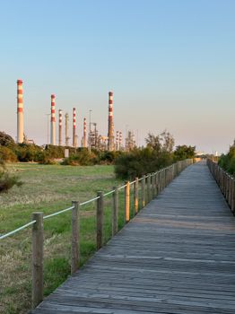 Wooden boardwalk beside grassy field and industrial smokestacks under the sunset sky.