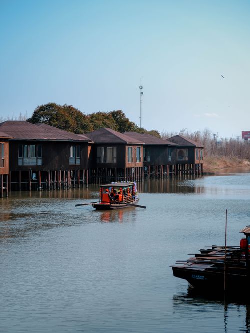 Free Traditional wooden stilt houses by a serene lake with a small boat passing by, under a clear blue sky. Stock Photo