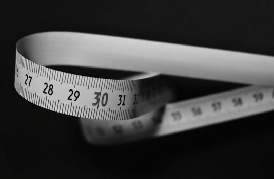 Black and white image of a flexible measuring tape curling on a dark background.