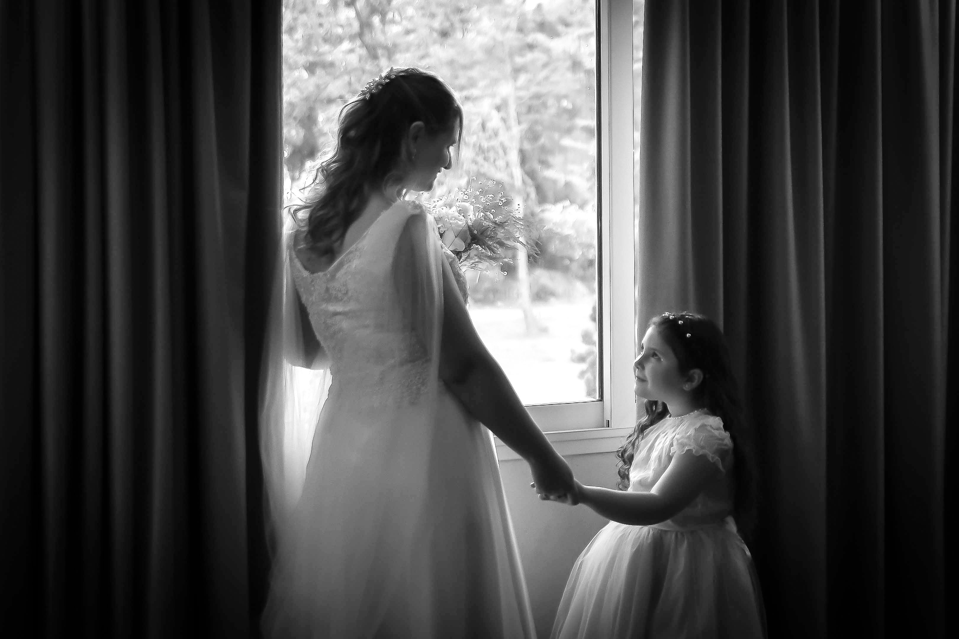 A beautiful black and white photo of a bride with a young girl near a window, filled with emotion.