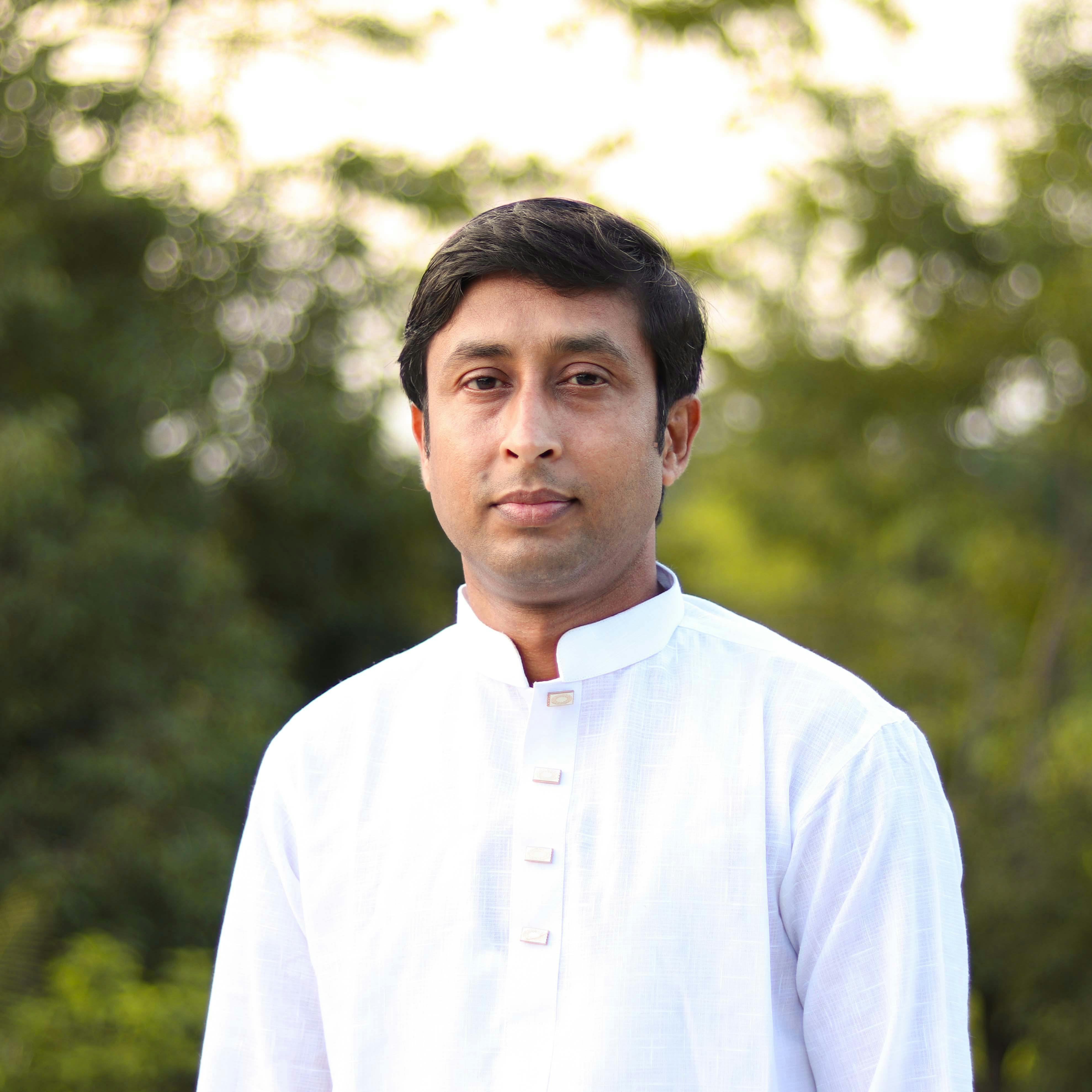 A serene portrait of an adult male outdoors in traditional white clothing, surrounded by lush greenery.