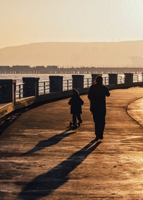 Free Silhouettes of a parent and child walking along a scenic waterfront path during the golden hour. Stock Photo