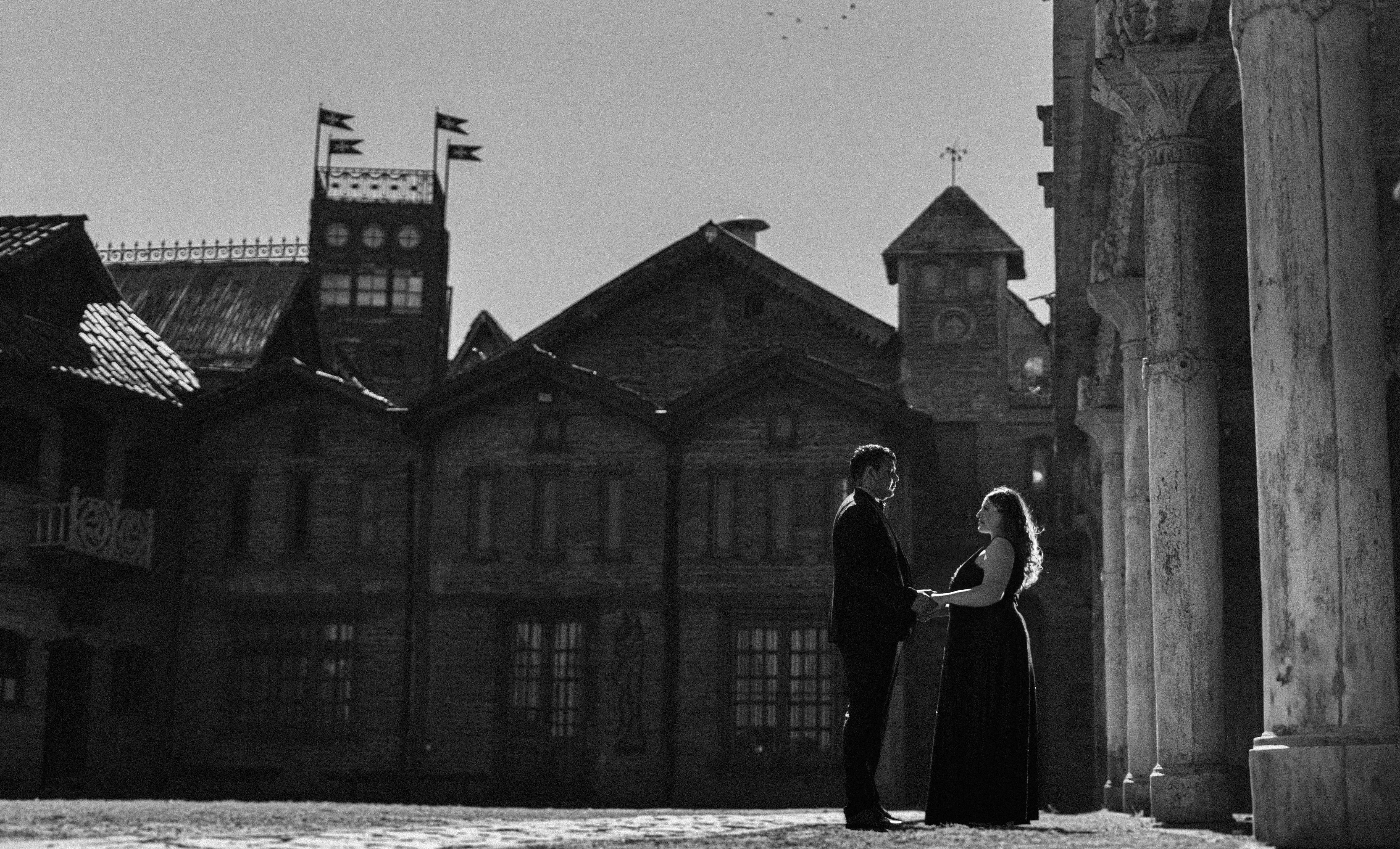 A couple stands holding hands in an elegant black and white setting, surrounded by historic architecture.