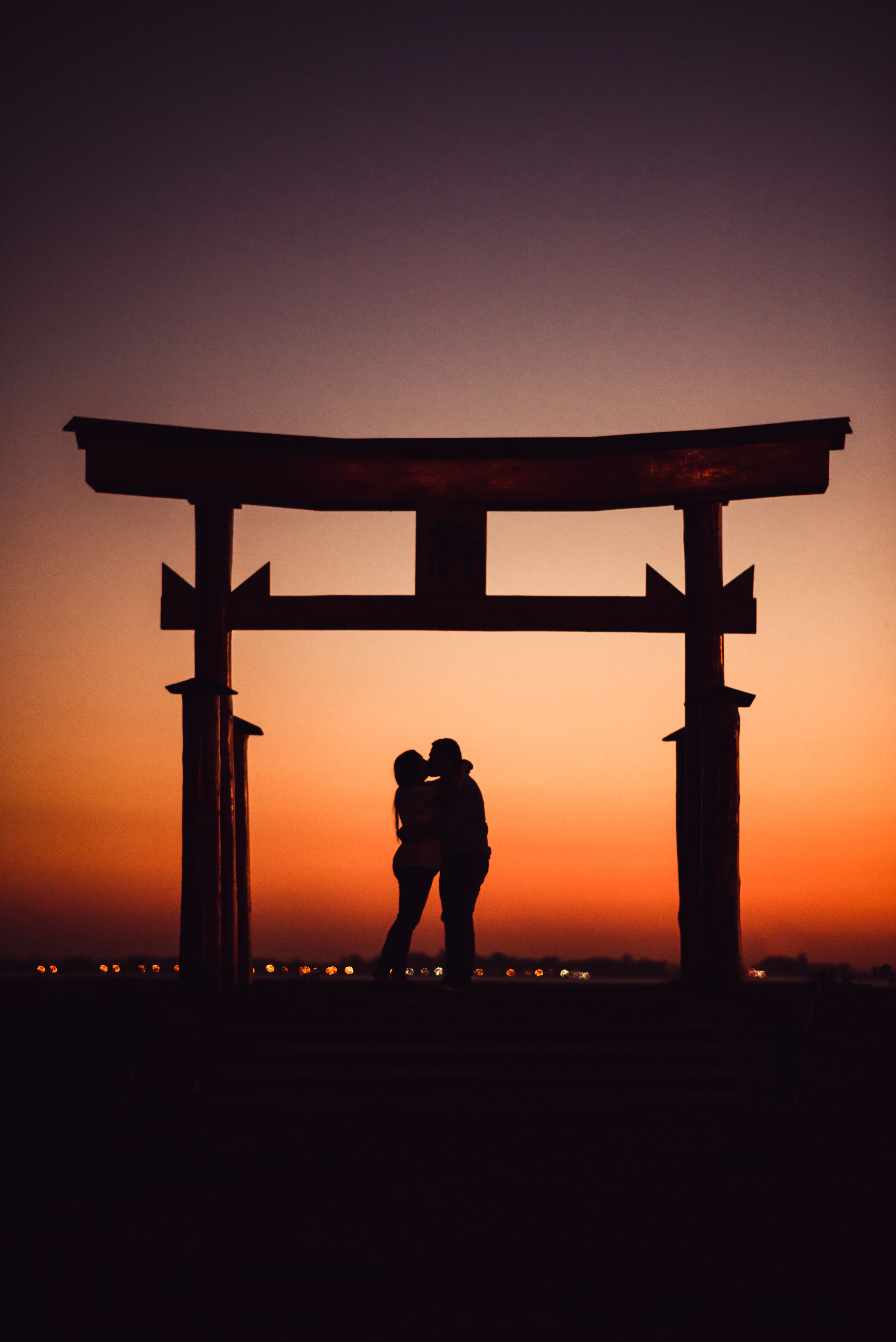 A romantic couple embraces in silhouette under a Torii gate at sunset, creating a serene moment.