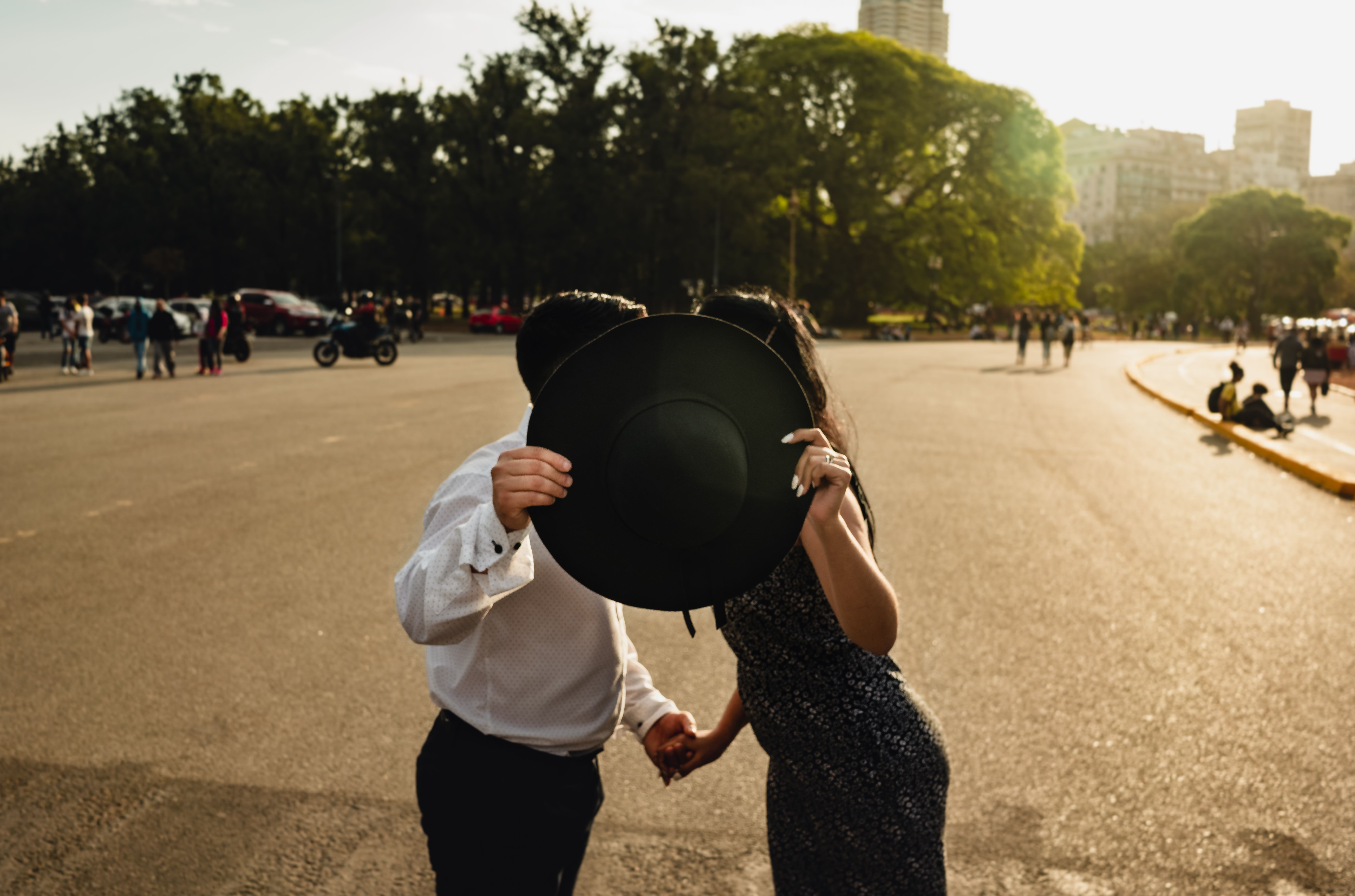 A couple playfully hides behind a hat while standing in a sunlit park, creating a romantic atmosphere.