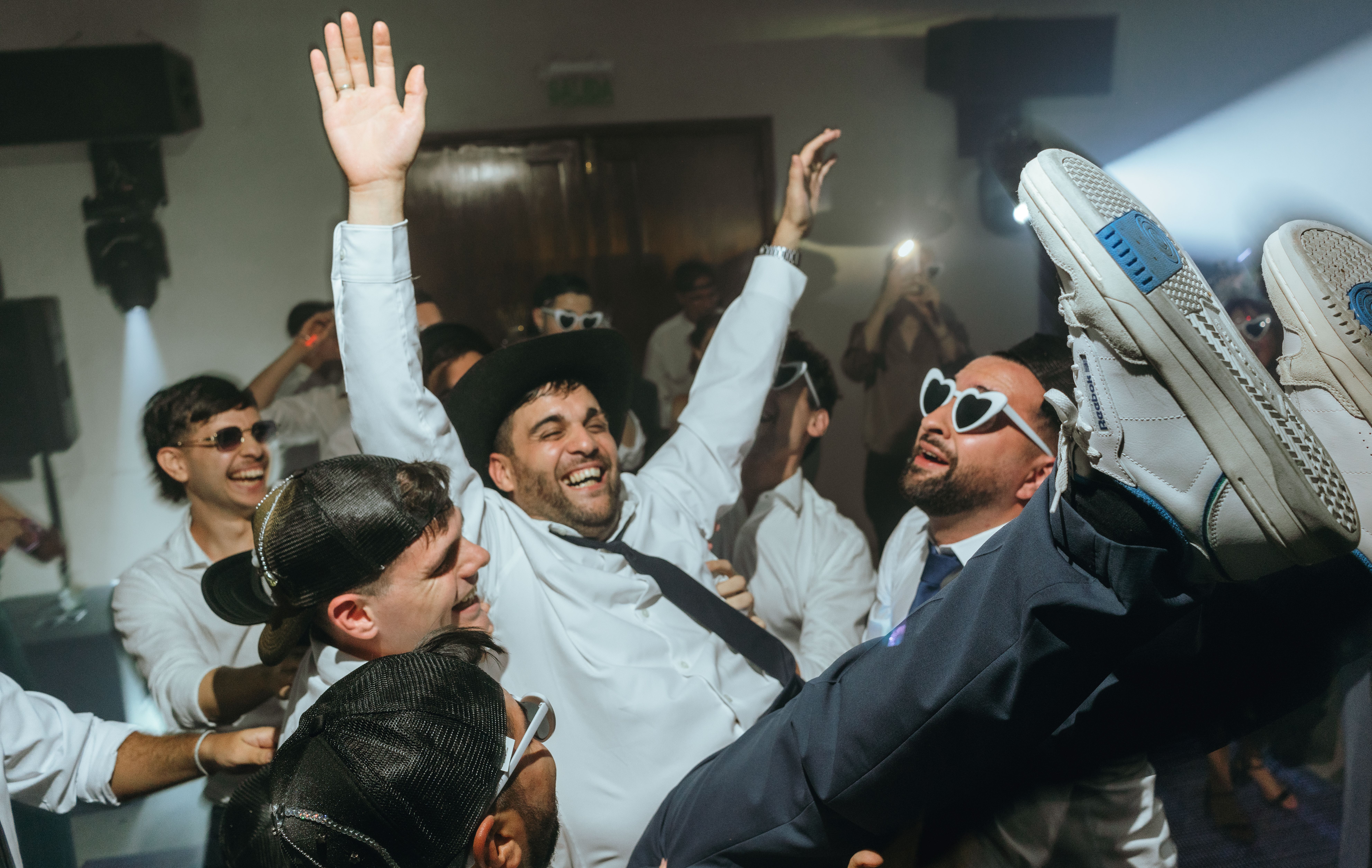 Group of friends lifting groom at a lively wedding reception. Joyful, festive atmosphere with heart-shaped sunglasses.