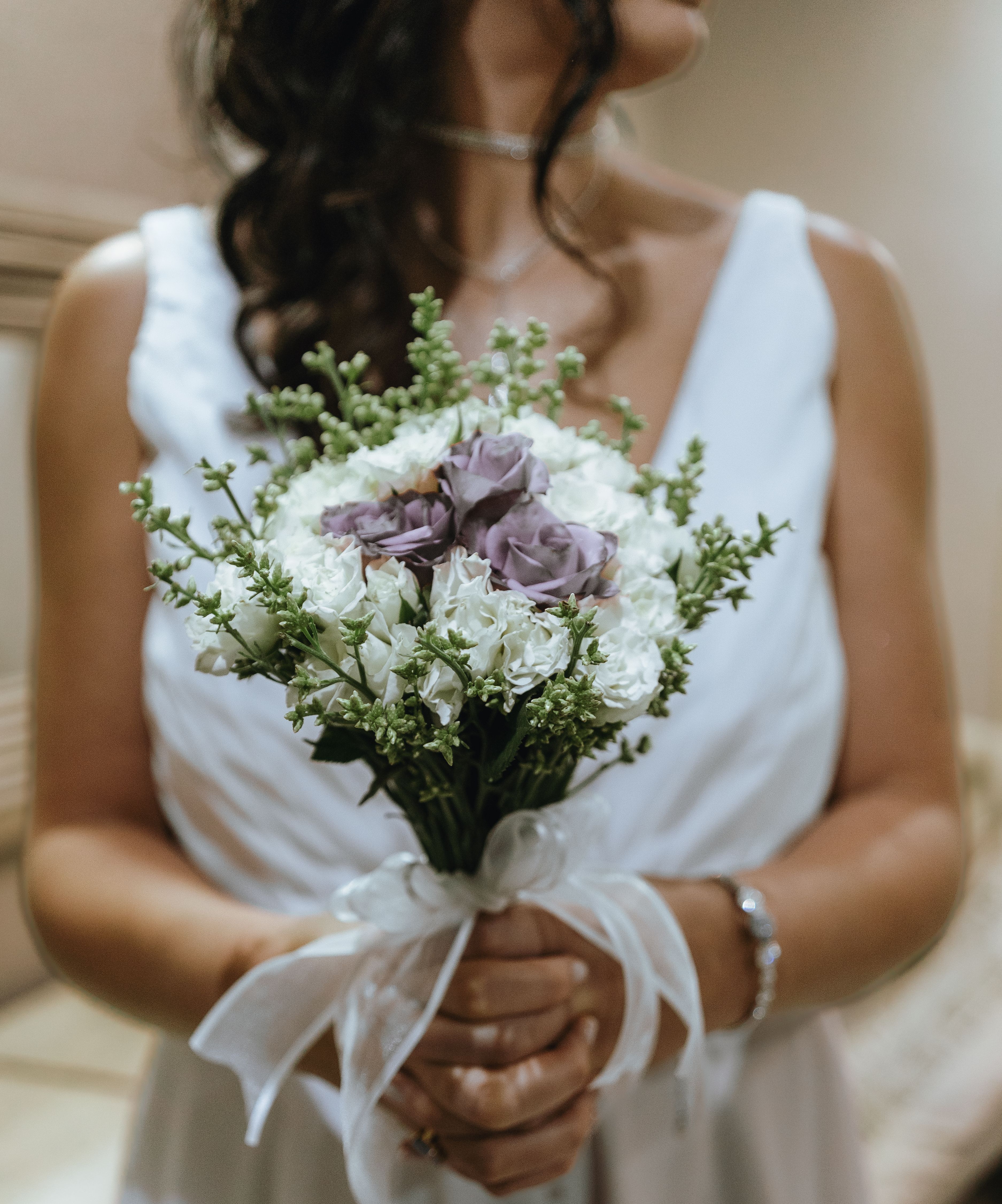 A bride in a white dress holds a beautifully arranged wedding bouquet indoors, creating an elegant and serene scene.