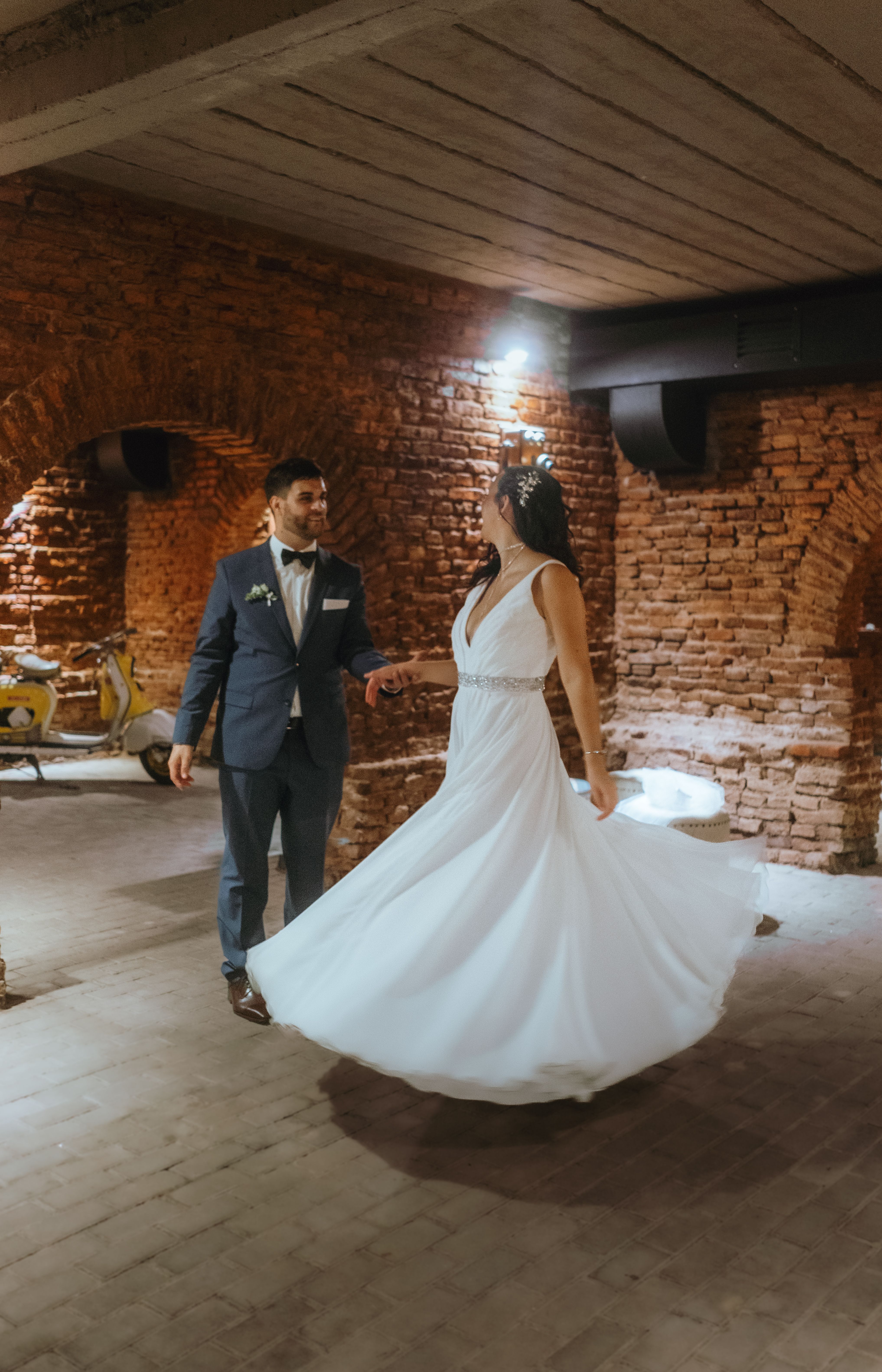 Bride and groom dancing in a rustic brick venue with elegant attire, capturing a romantic moment.