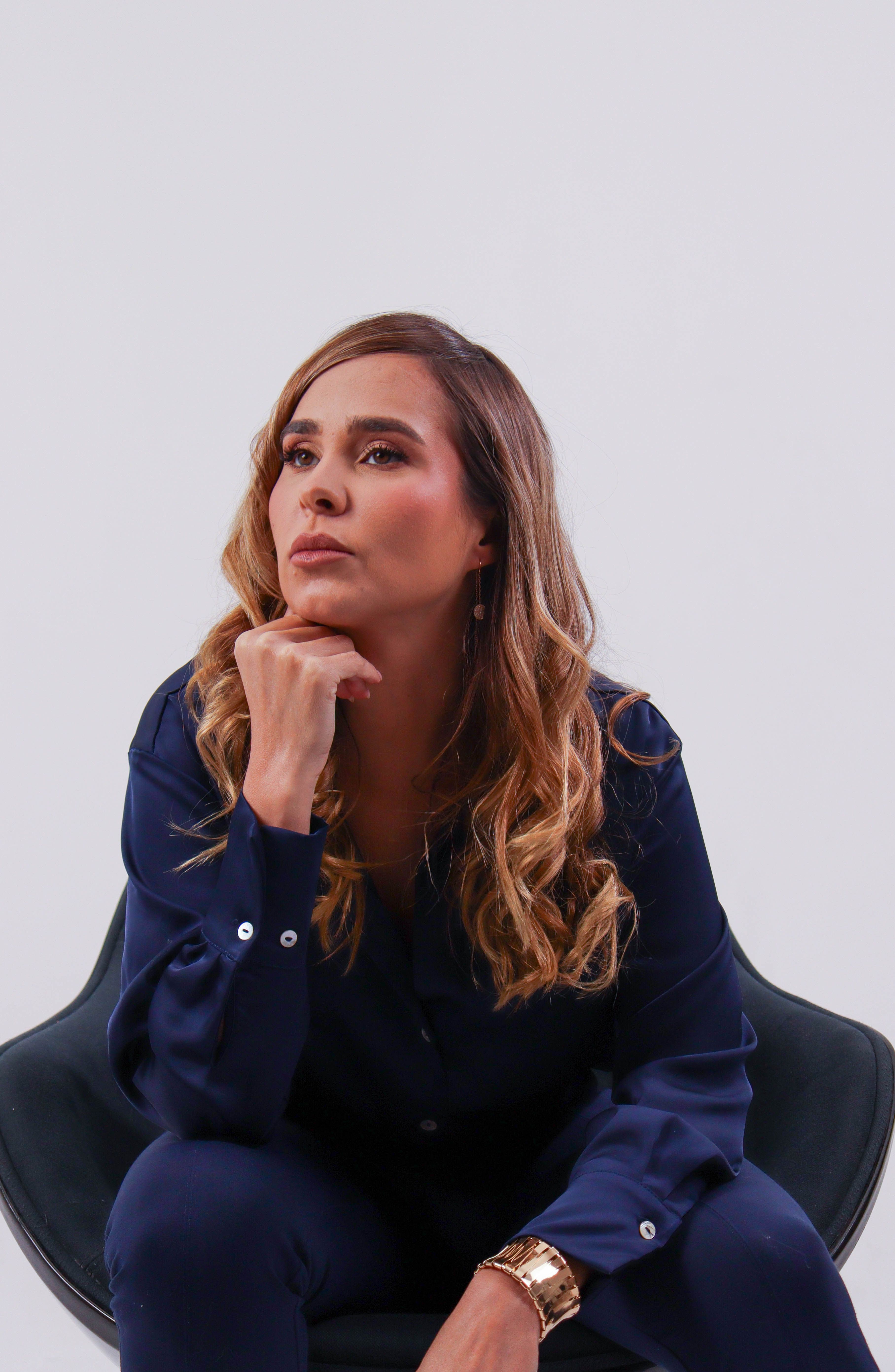 A thoughtful woman in a blue outfit sits on a chair, gazing into the distance.