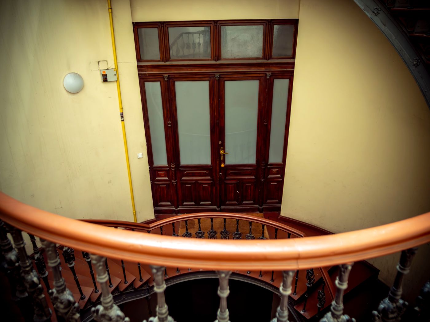 Cage d’escalier d’un immeuble ancien vue depuis un étage, main courante en bois verni, balustres tournés et porte vitrée en bois au rez-de-chaussée