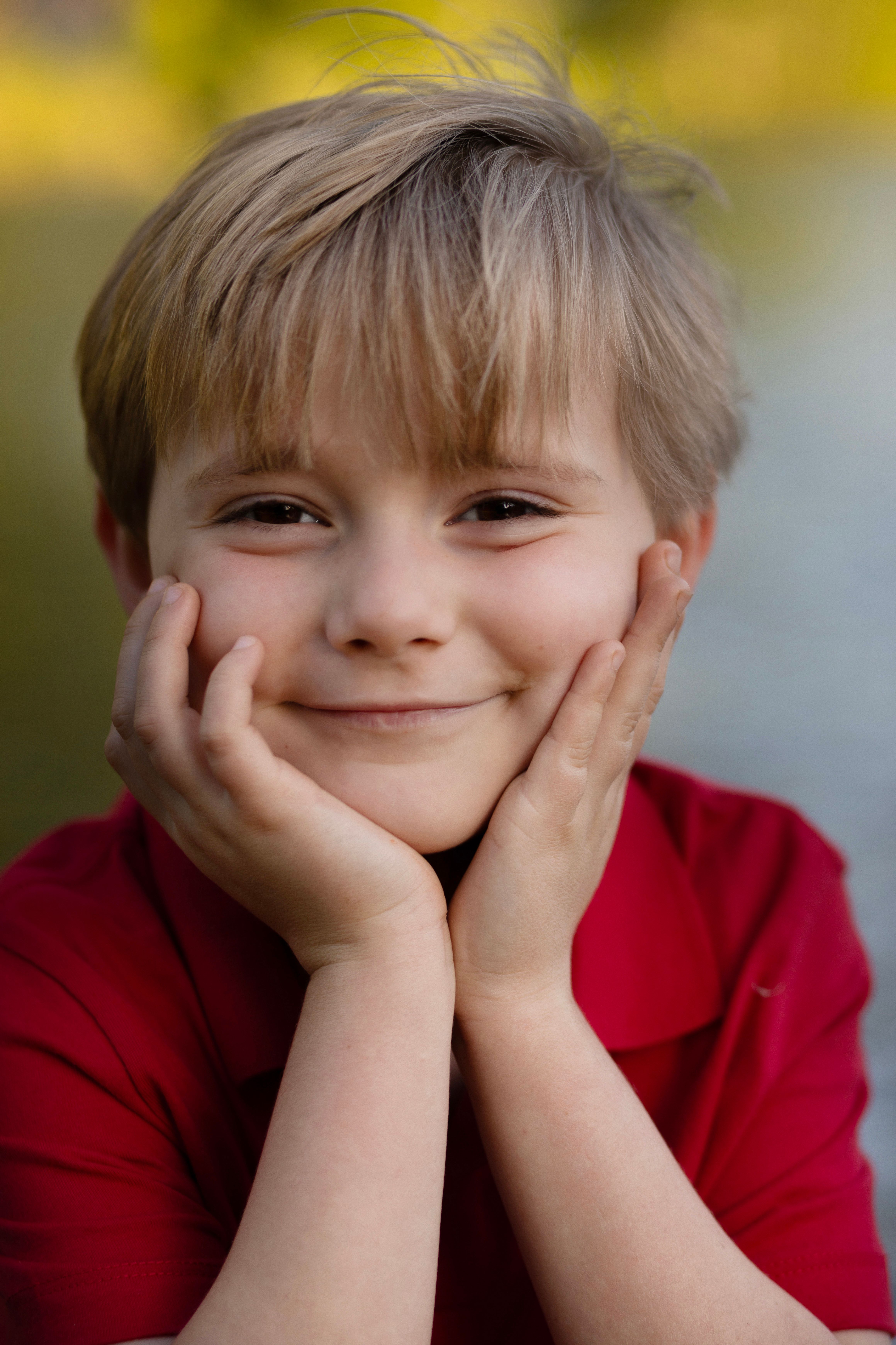 A young boy in a red shirt smiles warmly with hands on face outdoors.