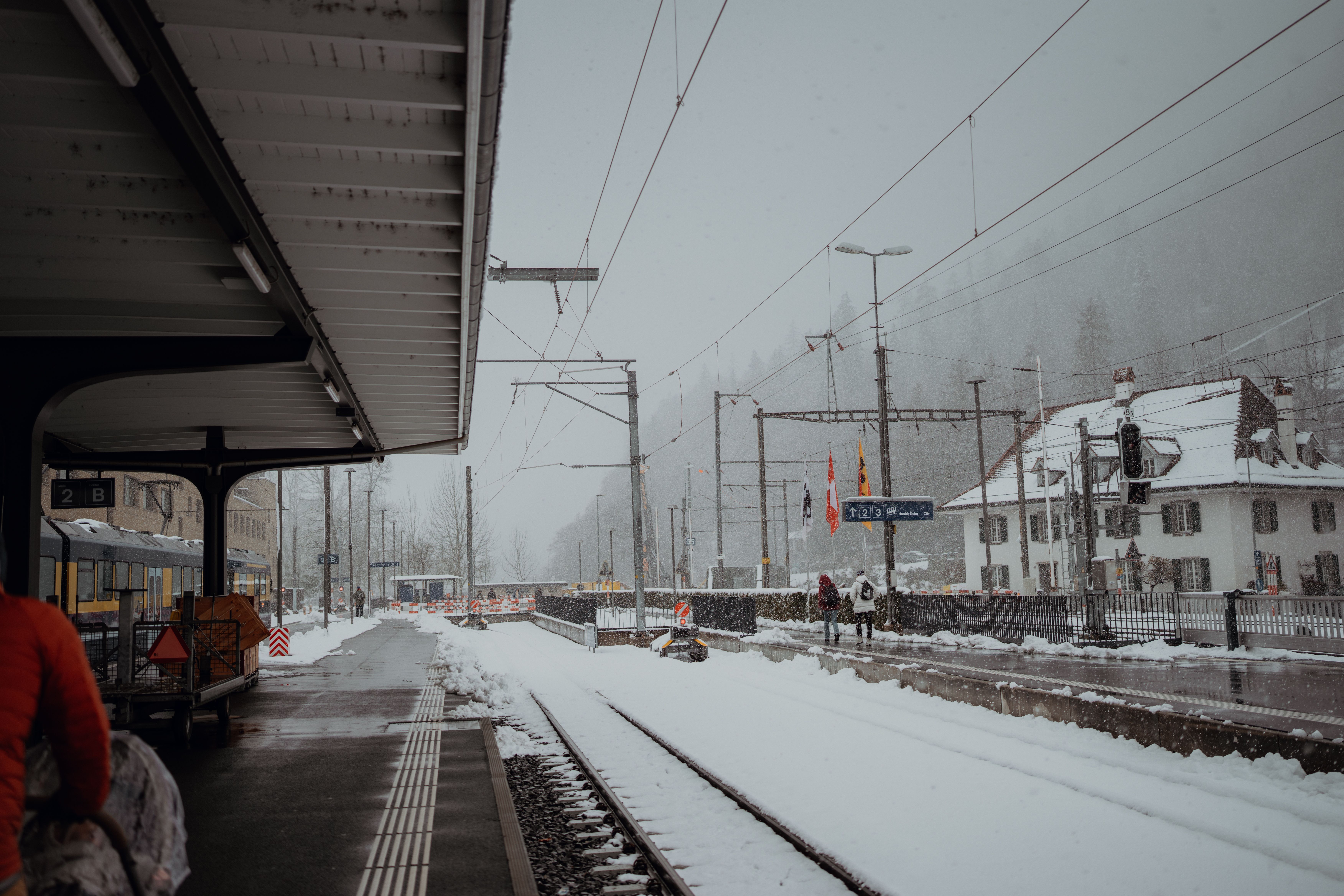 Travellers waiting with luggage on a snow-covered alpine railway platform while snow falls on the tracks