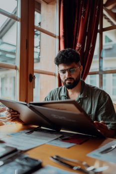 Man wearing glasses intently reading a menu at a cozy indoor cafe setting, surrounded by paperwork.