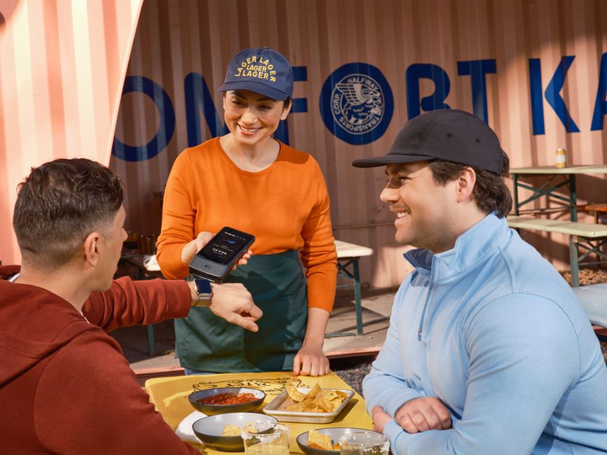 Friends using smartwatch for contactless payment at an outdoor cafe, assisted by a smiling waitress. Friends using smartwatch for contactless payment at an outdoor cafe, assisted by a smiling waitress.