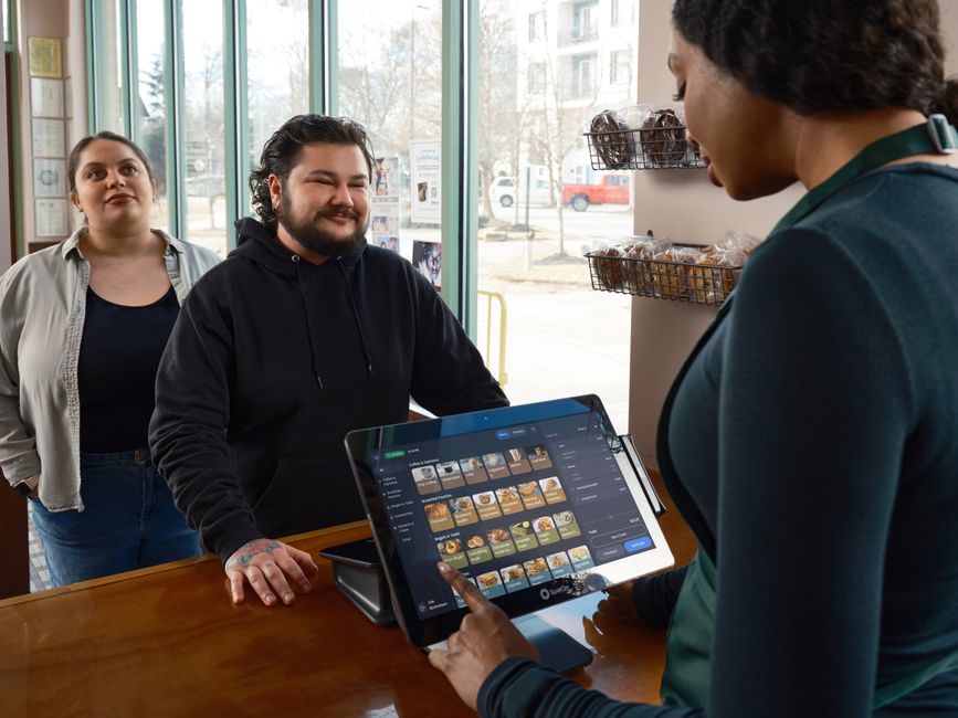 Same Day Merchant Cash Advance: Funding In 24 Hours 1 A barista stands behind a counter taking an order on a touchscreen register—part of a café supported by merchant cash advance—while two customers stand on the other side, smiling and waiting in the bright space.