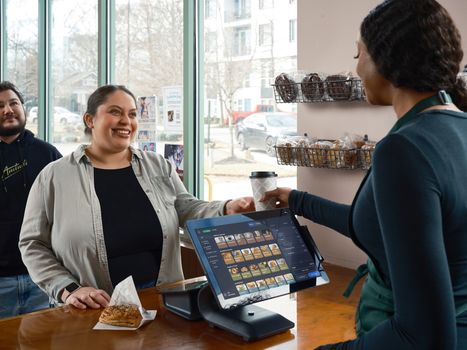 Barista uses modern POS system in café; smiling customer with pastry and coffee in hand. Indoor café setting.