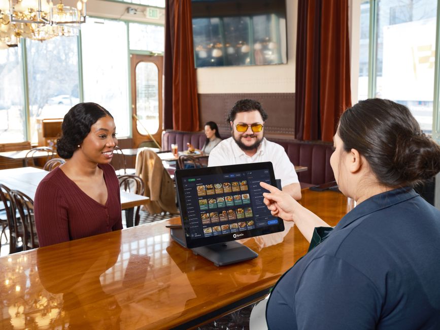 Server demonstrating a SpotOn POS system for customers in a cozy restaurant setting. Server demonstrating a SpotOn POS system for customers in a cozy restaurant setting.
