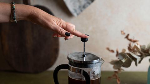 A hand presses a French press with fresh coffee on a wooden surface in a cozy kitchen setting.