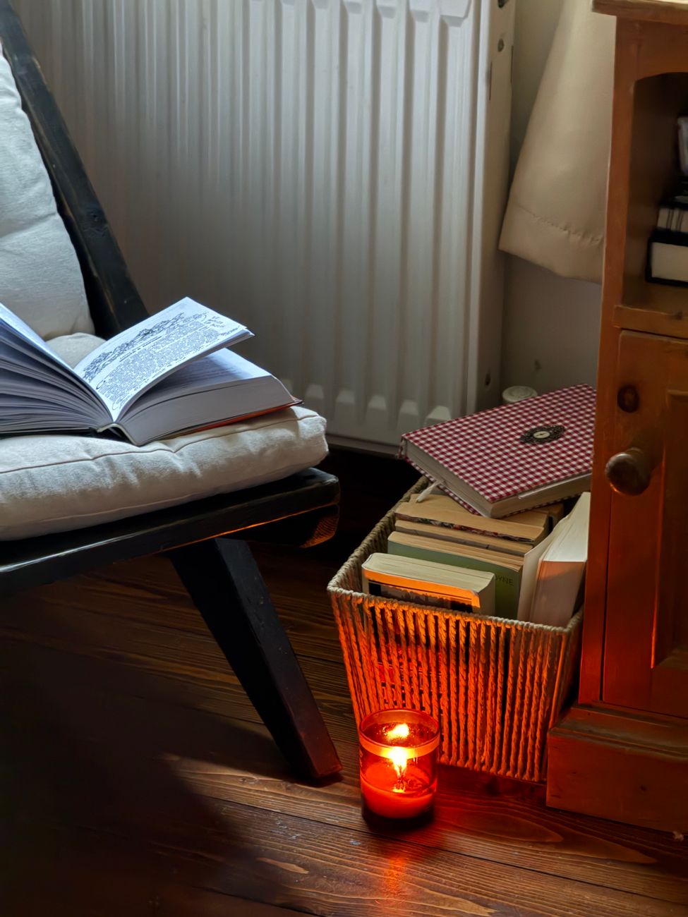A warm indoor scene featuring an open book, soft chair, basket of books, and a lit candle, creating a relaxing atmosphere.