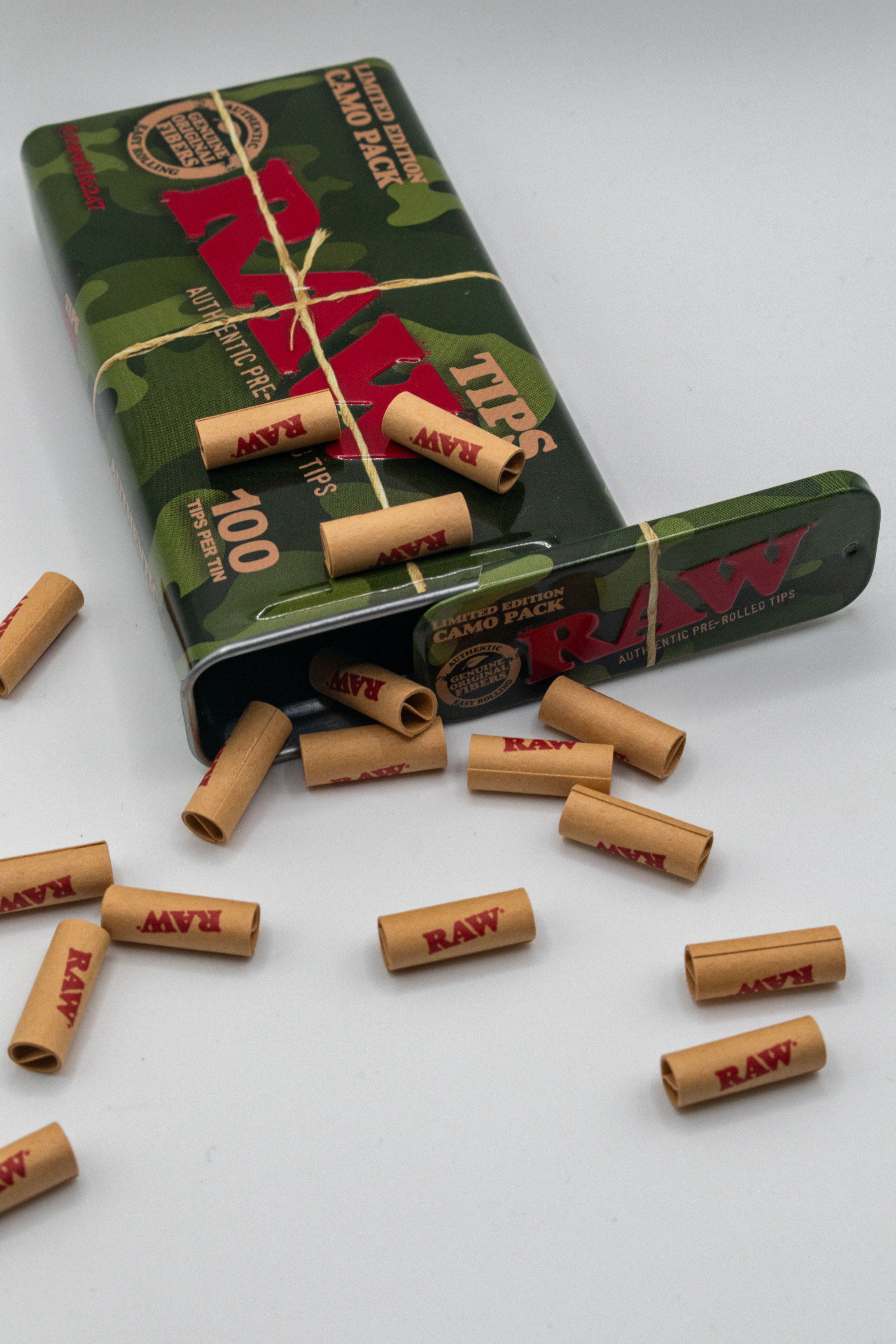 Colorful RAW organic rolling tips spill from a camouflage tin box against a white background.
