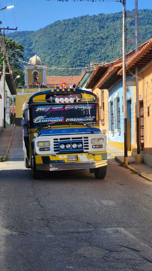 Free Vibrant bus navigates a quaint street under a clear blue sky with mountains in the background. Stock Photo