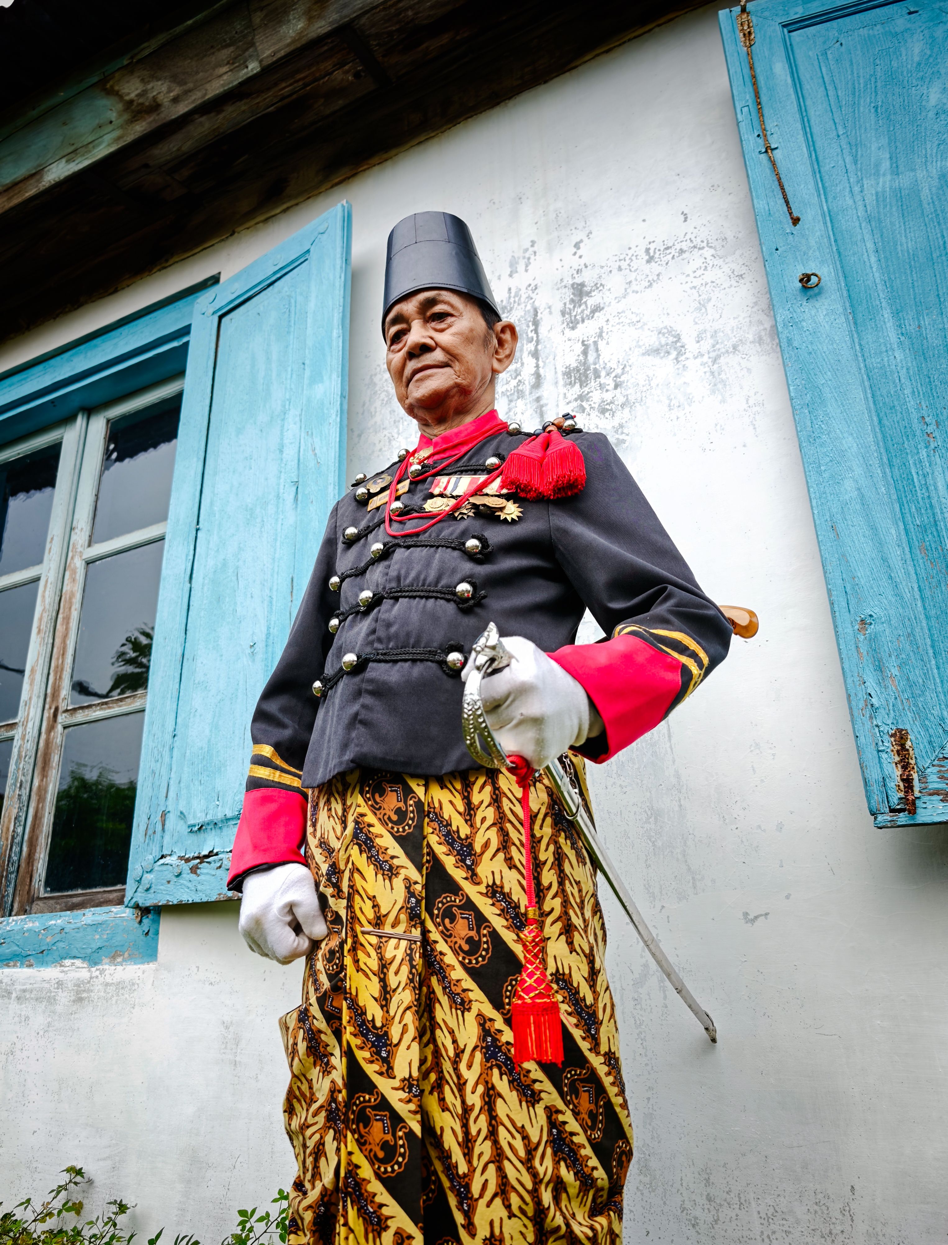 Senior man wearing traditional Indonesian clothing stands by wooden house with blue shutters.
