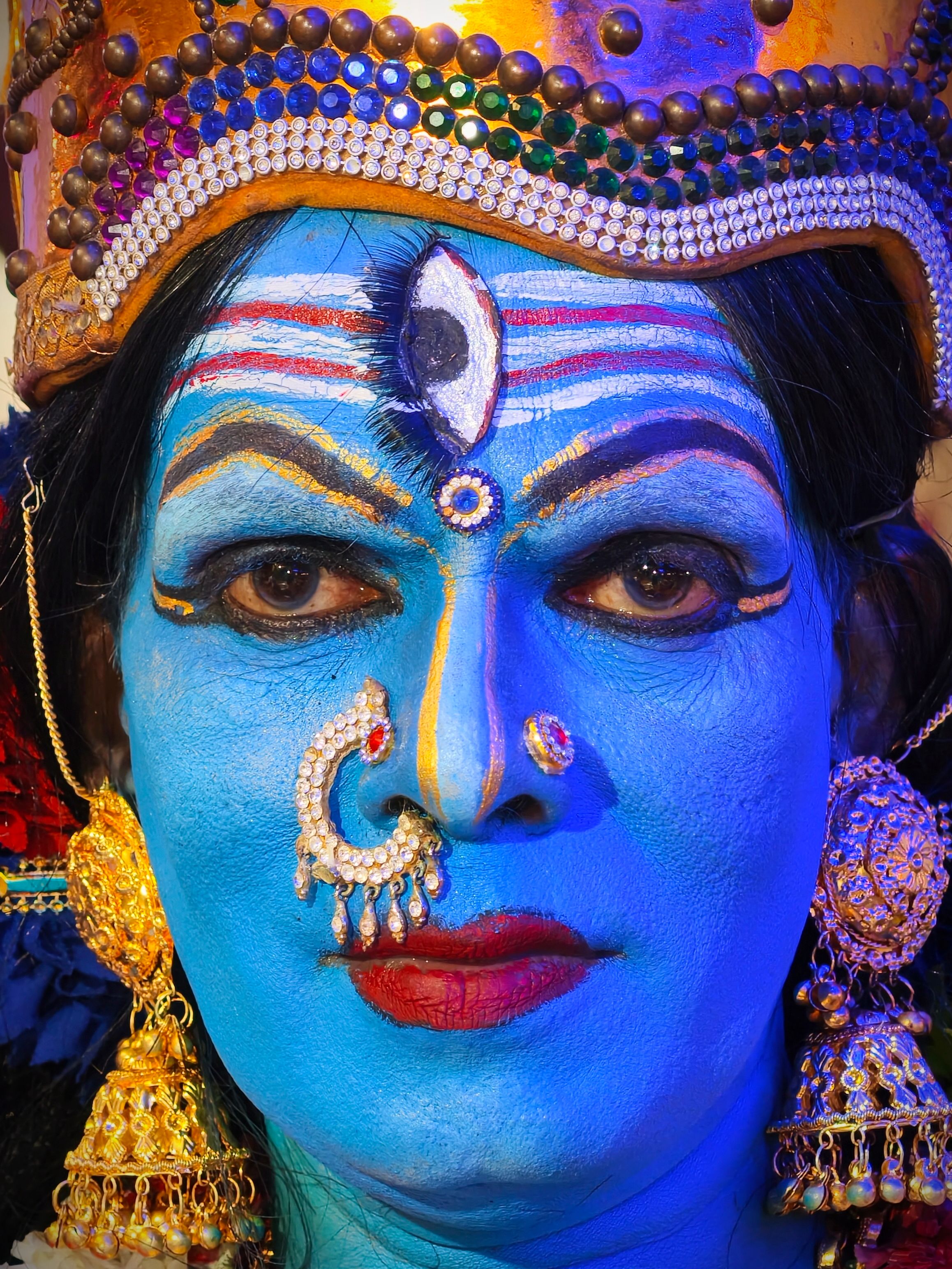 Close-up of a colorful traditional Indian face paint adorned with intricate jewelry and a gold headdress.