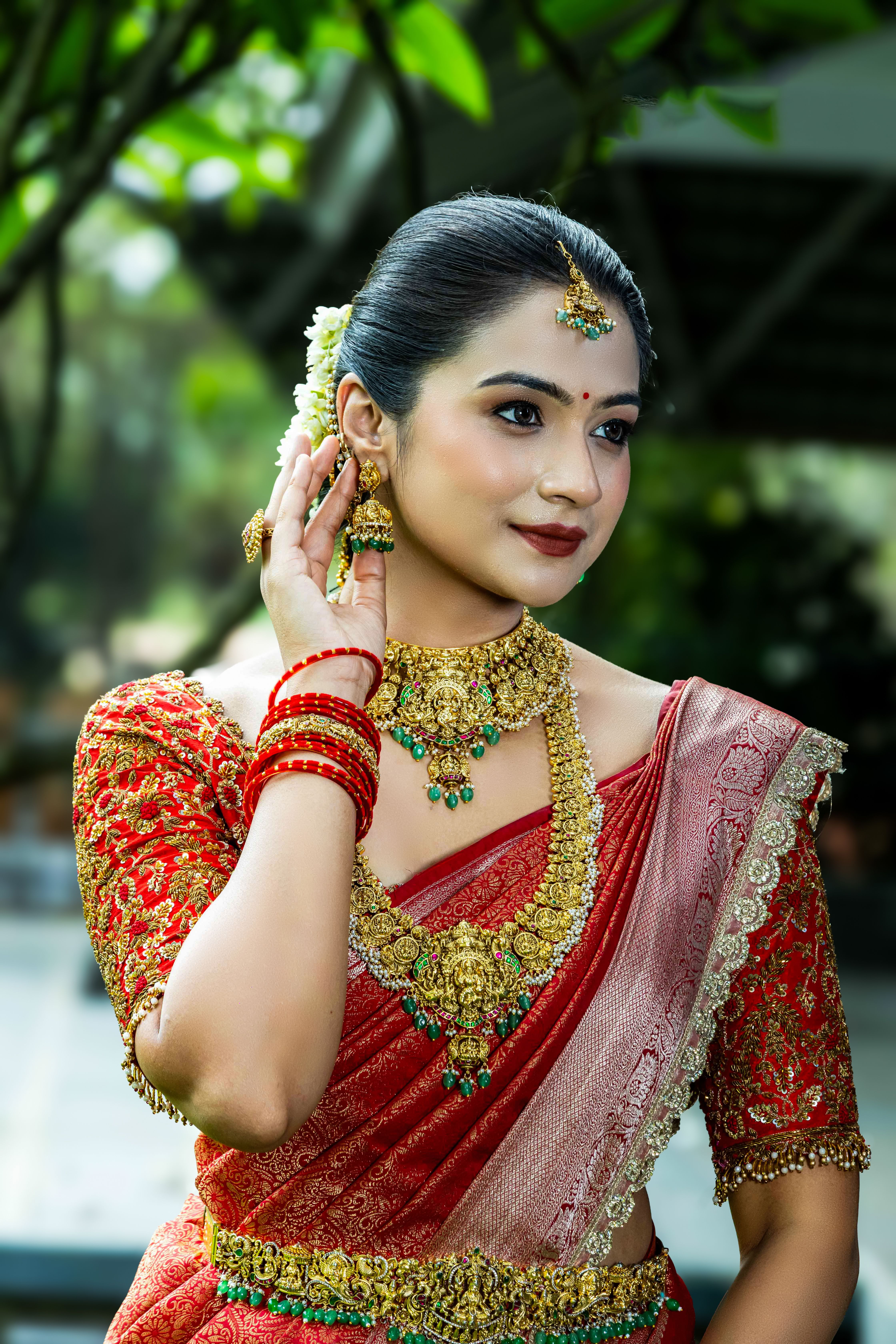 Elegant Indian bride in traditional red and gold attire with ornate jewelry, outdoors setting.