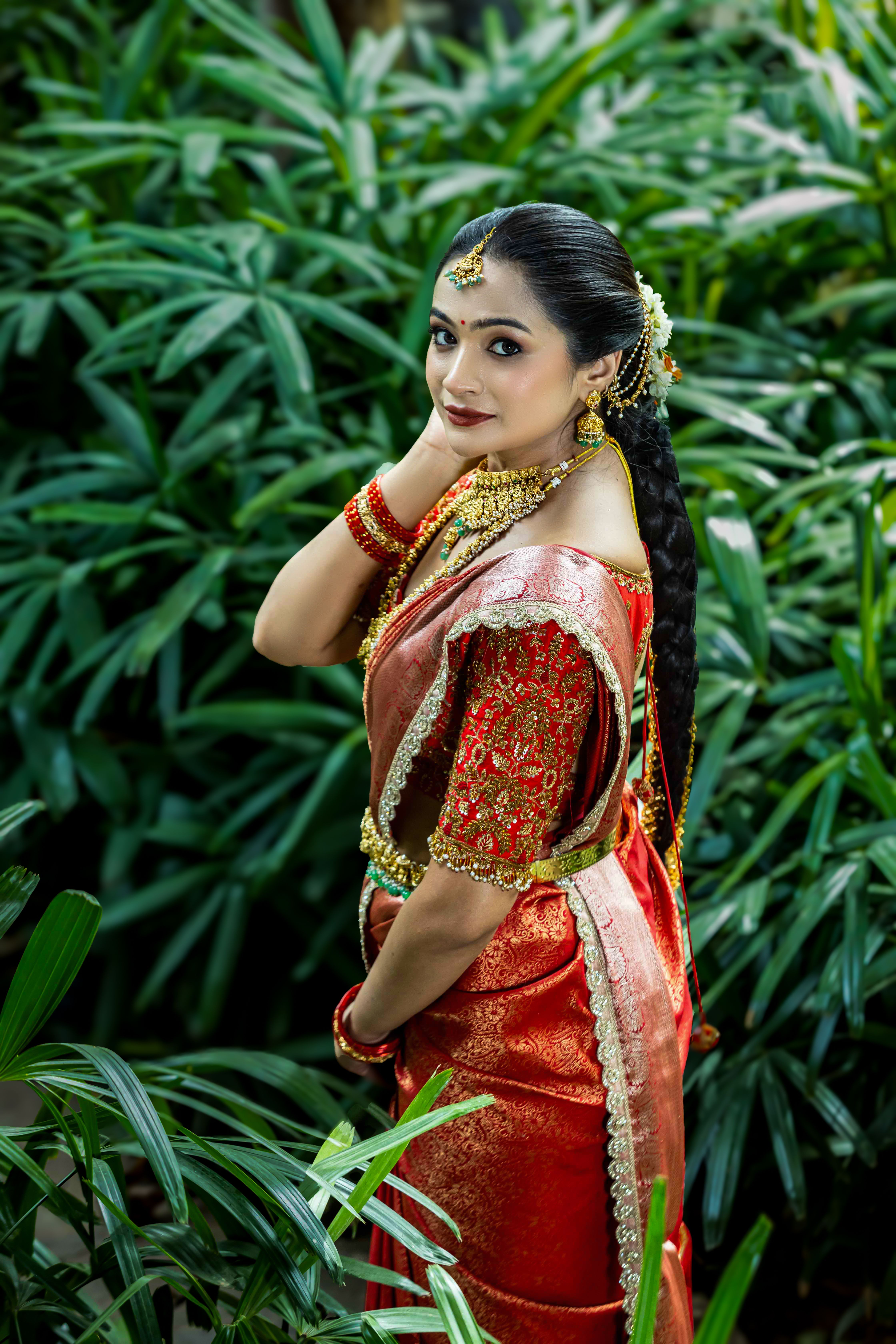 Graceful woman in a red silk saree, adorned with gold jewelry, surrounded by lush greenery.