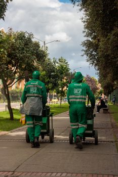 Two street cleaners in Bogotá pushing carts on a tree-lined sidewalk.