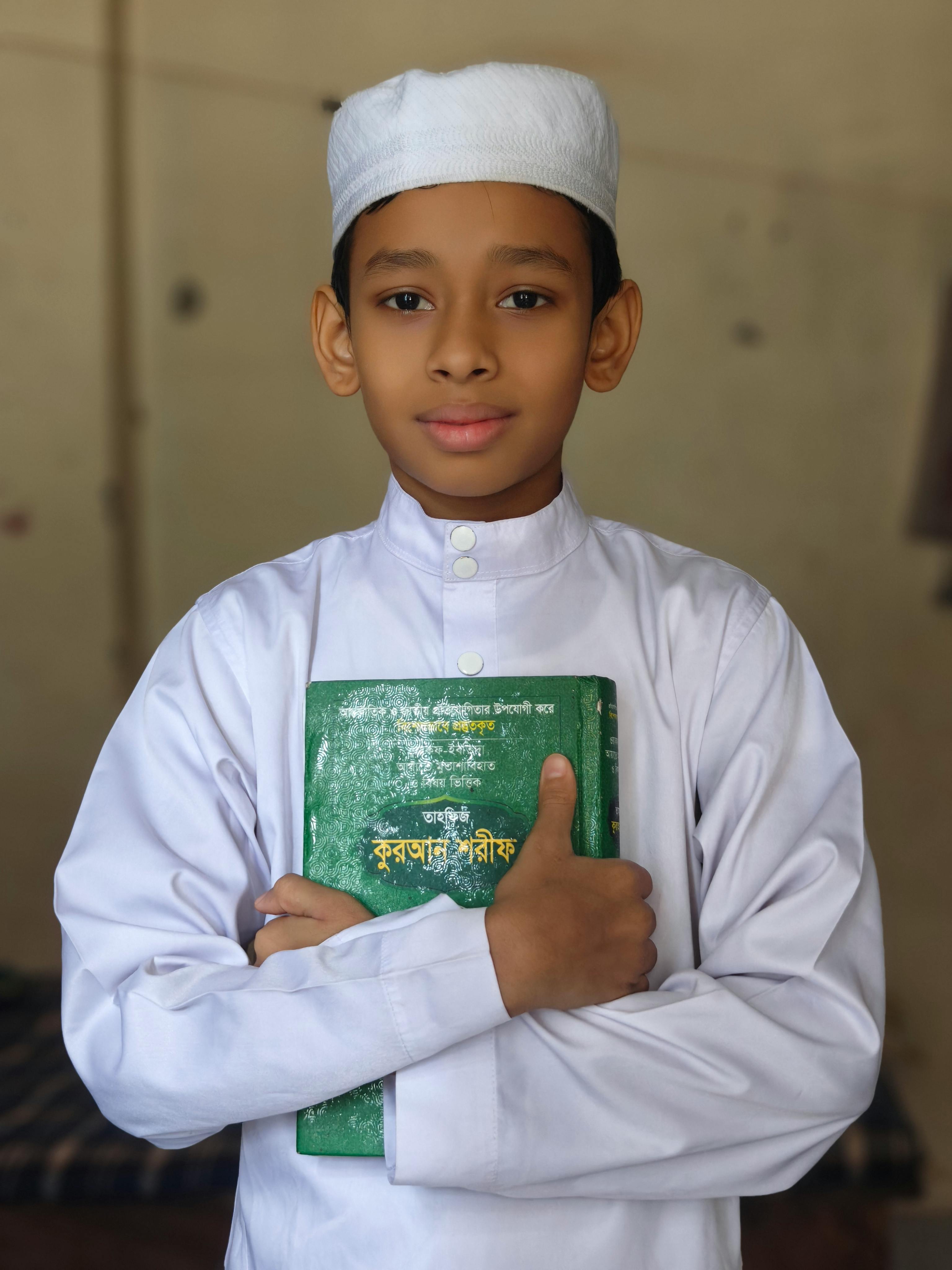 A young boy in traditional Muslim attire holding a green book indoors.