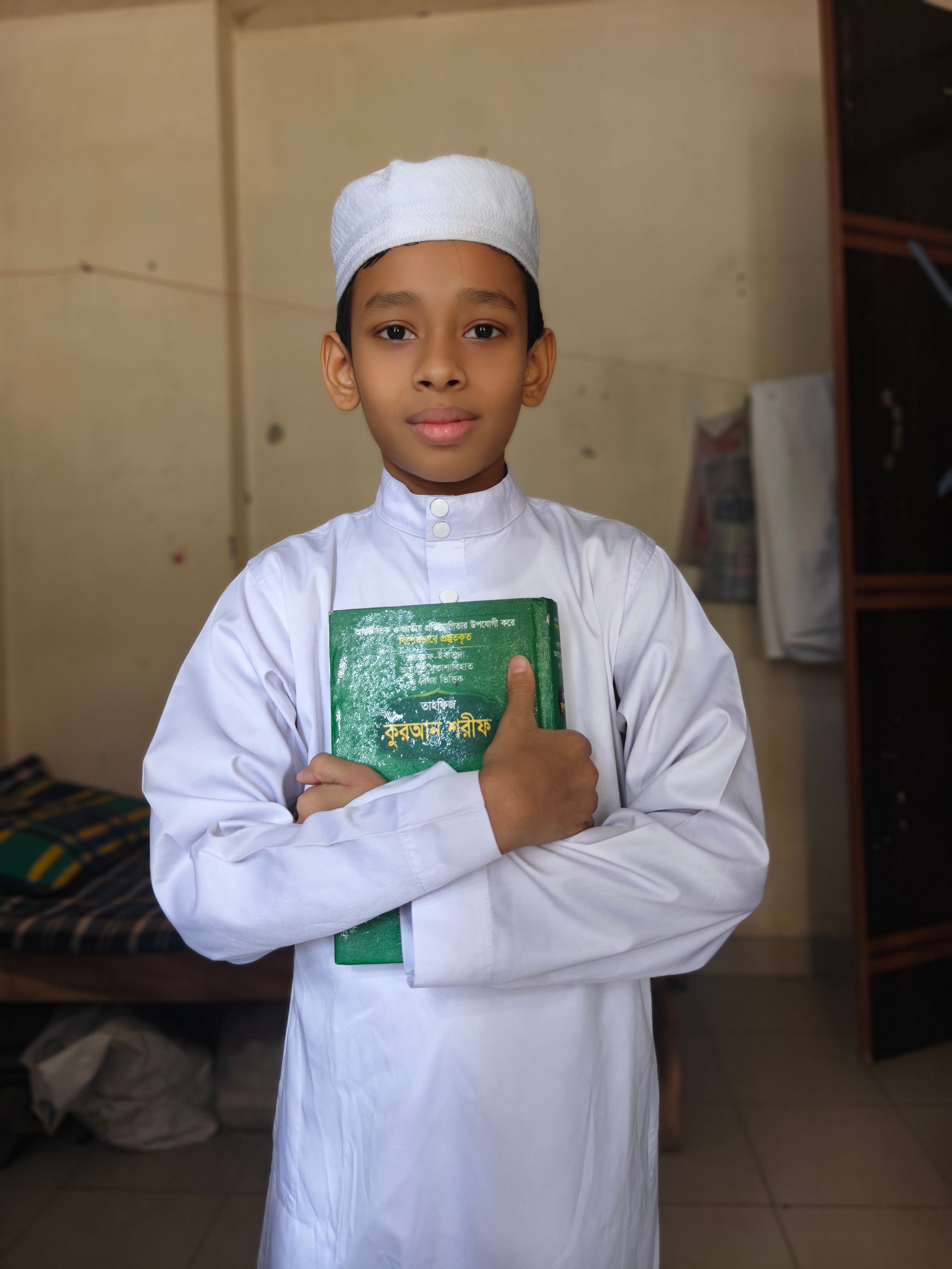 Youthful boy in white traditional attire, holding a green book, standing indoors with a calm expression.