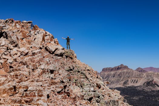 A climber celebrates atop a rugged mountain peak under a clear blue sky.