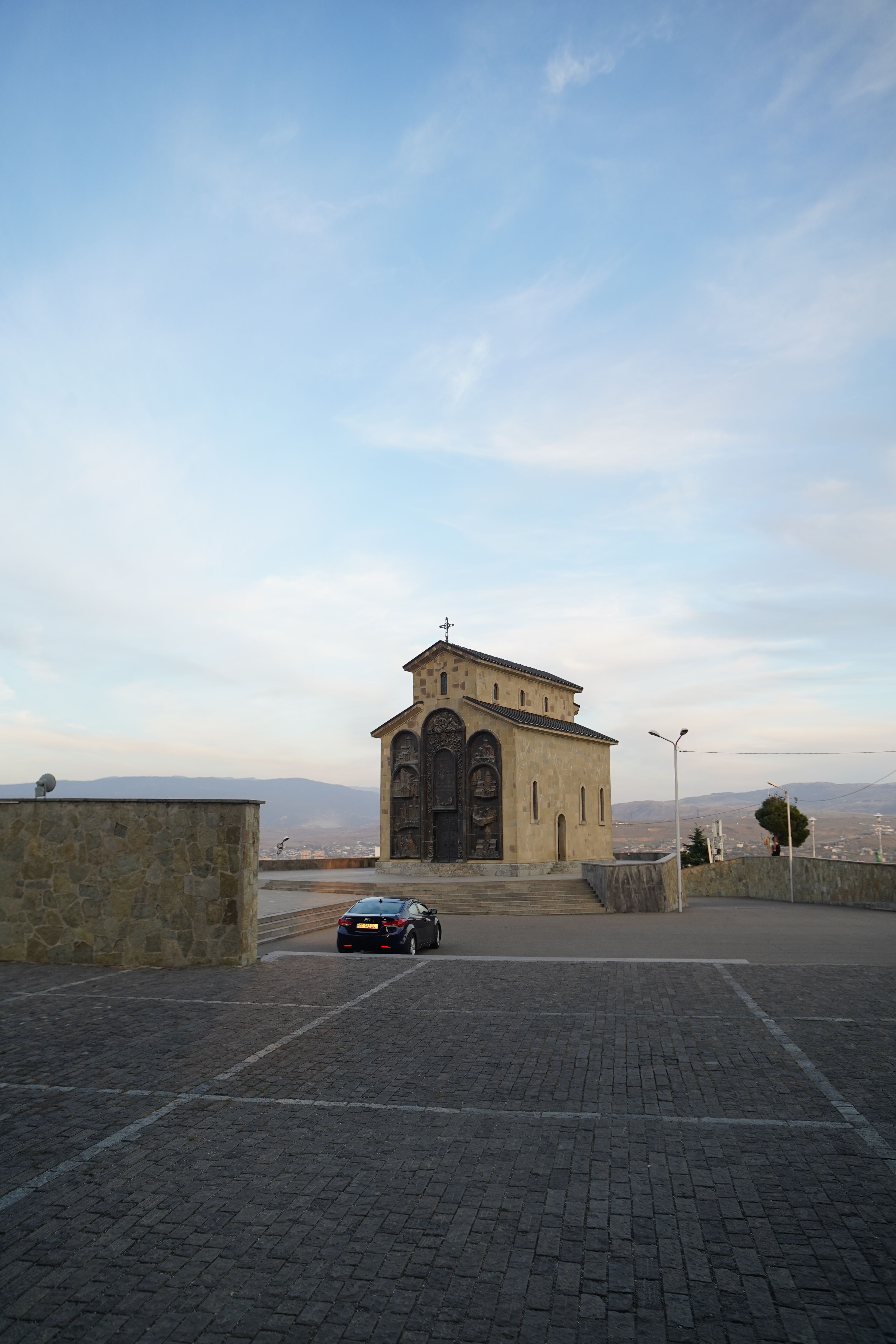 A scenic view of a historic church with a parked car and open sky in Tbilisi, Georgia.