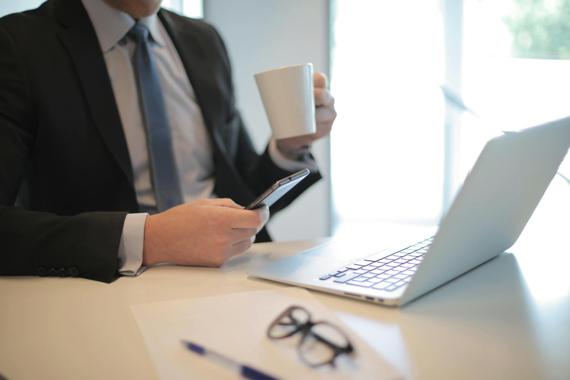 A businessman in a black suit working at a modern office desk with a laptop, phone, and coffee.