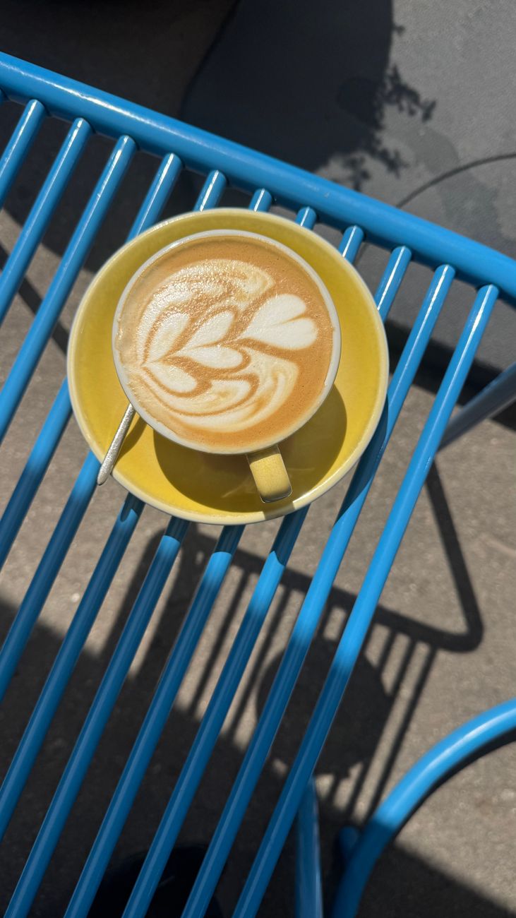 Artistic latte in a yellow cup on a bright blue metal table casting shadows in sunlight.