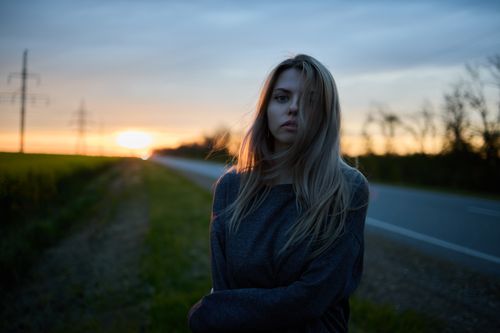 Free Portrait of a young woman standing by a rural road at sunset, gazing into the distance. Stock Photo