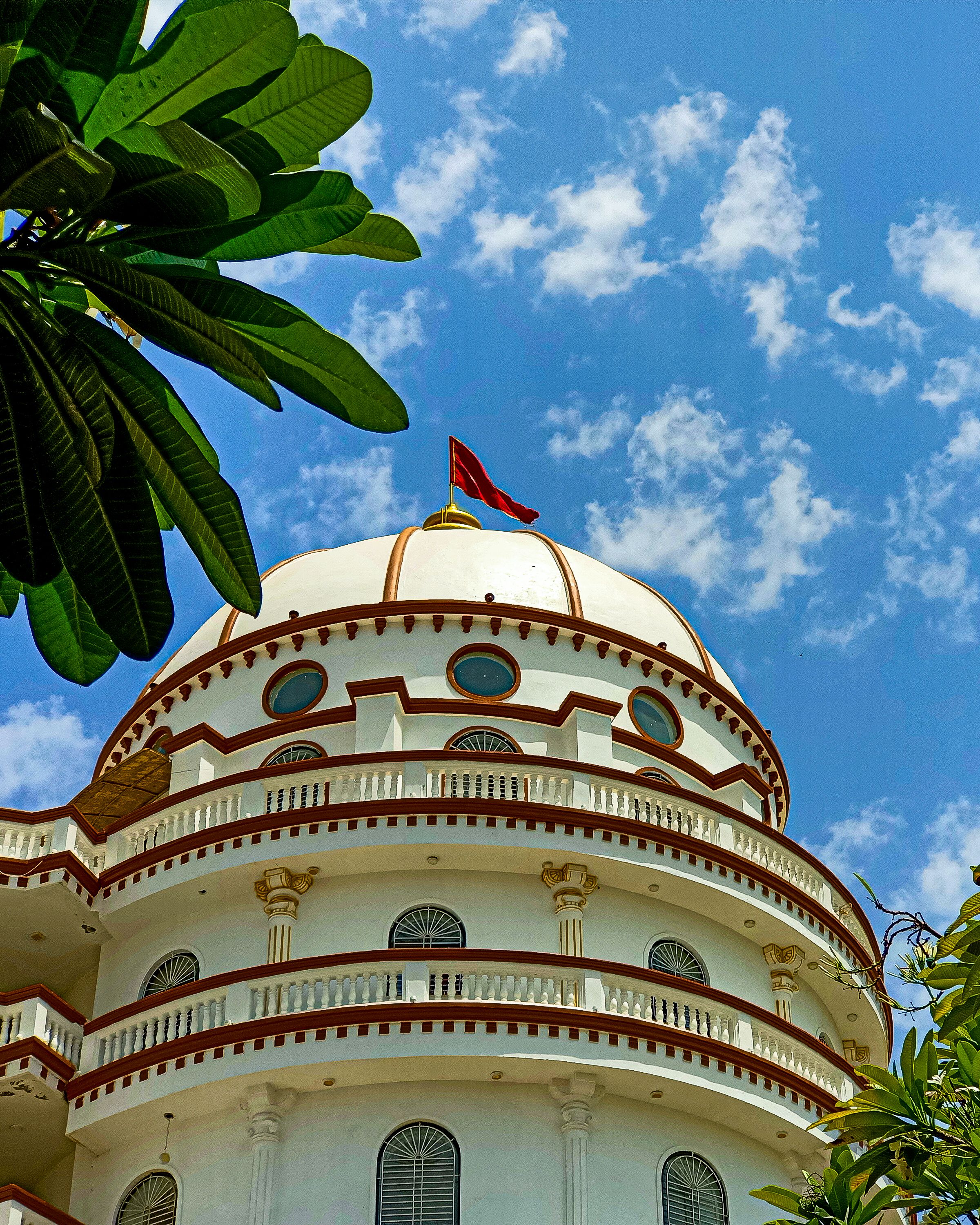 A beautiful white dome building with a flag against a bright blue sky, framed by lush green leaves.