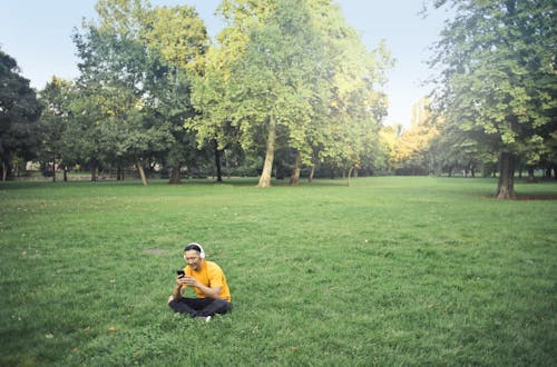 Photo of Man in Yellow T-shirt and Black Pants Sitting on Green Grass Field While Listening to Music on Headphones and Using His Phone