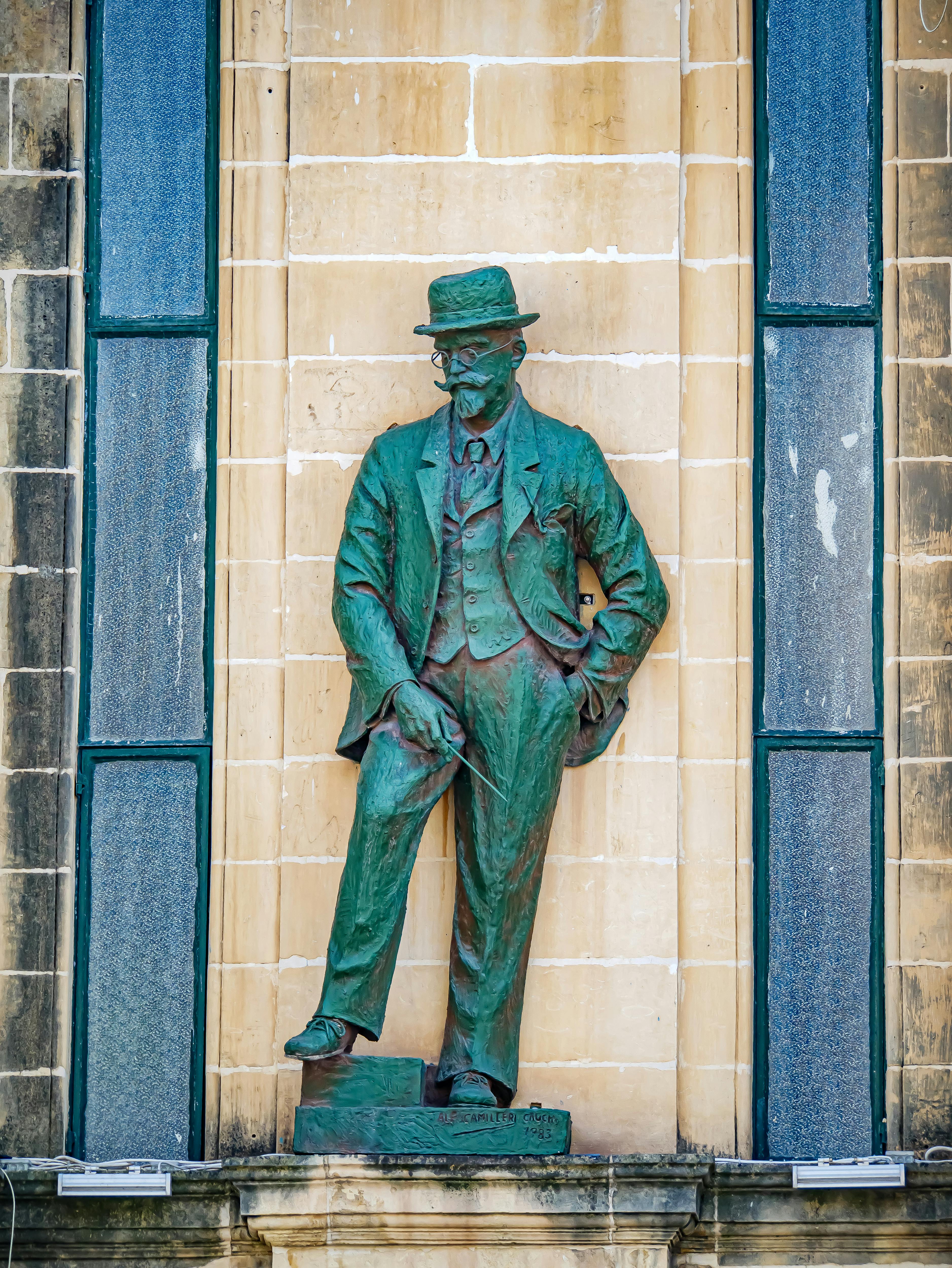 Bronze statue of a gentleman in a suit and hat on an ornate stone wall with green accents.