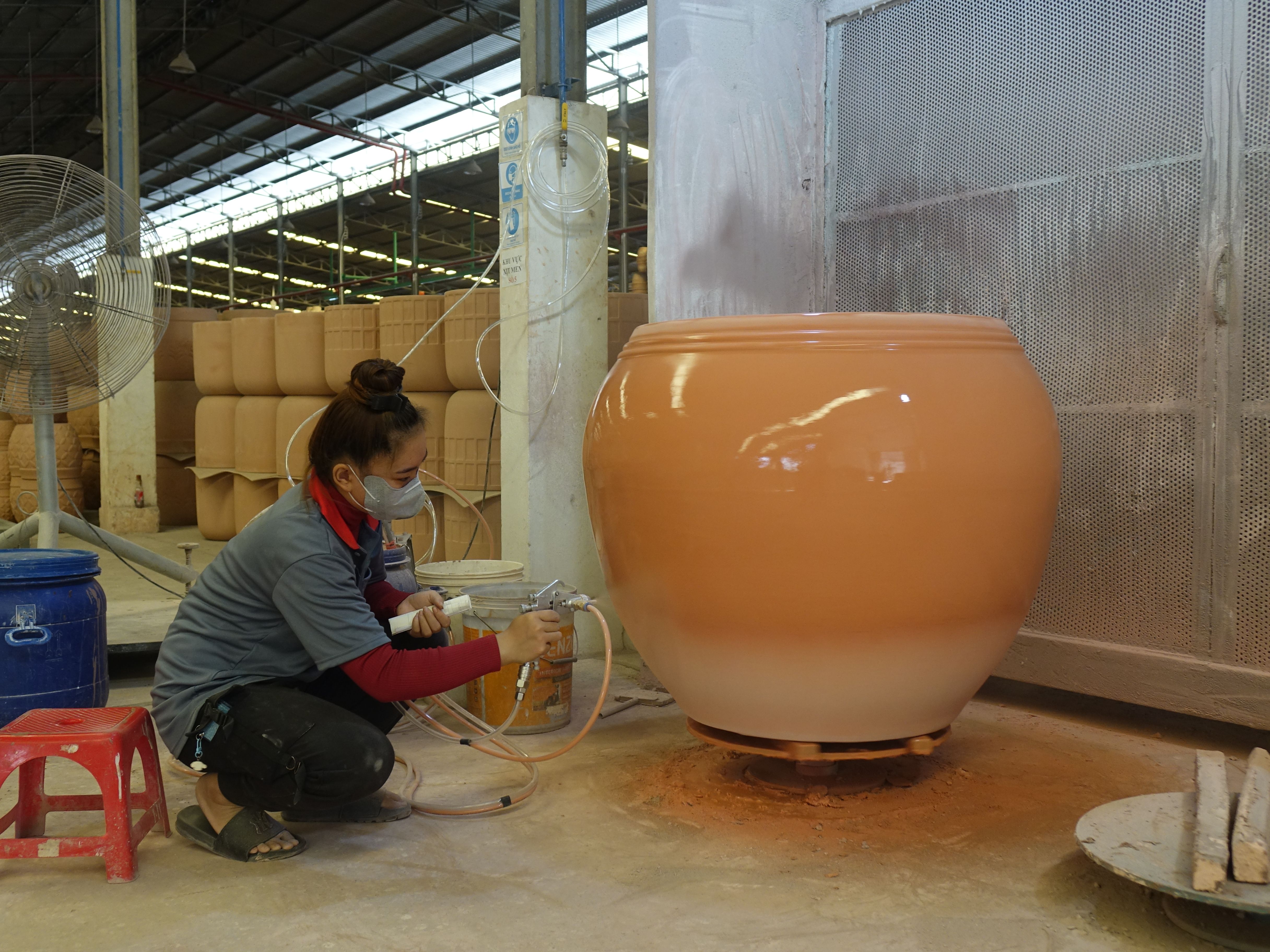 A worker diligently spray paints a large ceramic pot in a factory setting, adding a glossy finish.