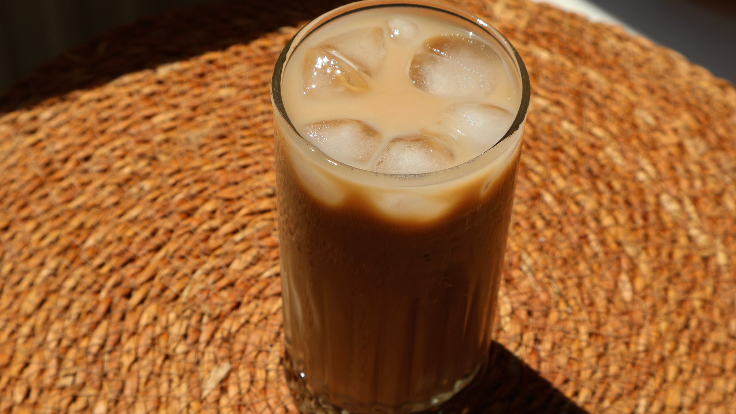A glass of iced coffee with ice cubes on a woven mat in bright sunlight.