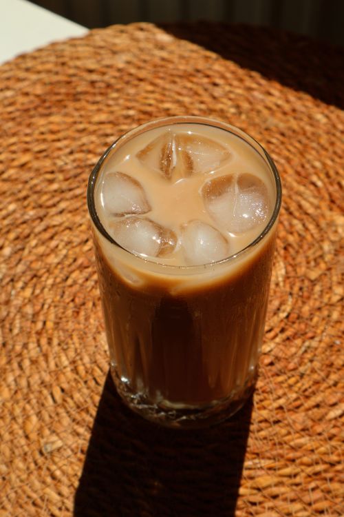 Free A tall glass of iced coffee with ice cubes on a woven placemat, sunlight casting a distinct shadow. Stock Photo